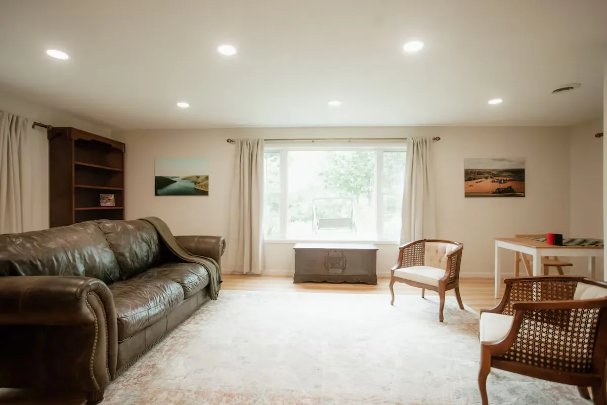 Living room with a leather sofa, two armchairs, a bookshelf, paintings on the wall, a window, and a wooden chest.
