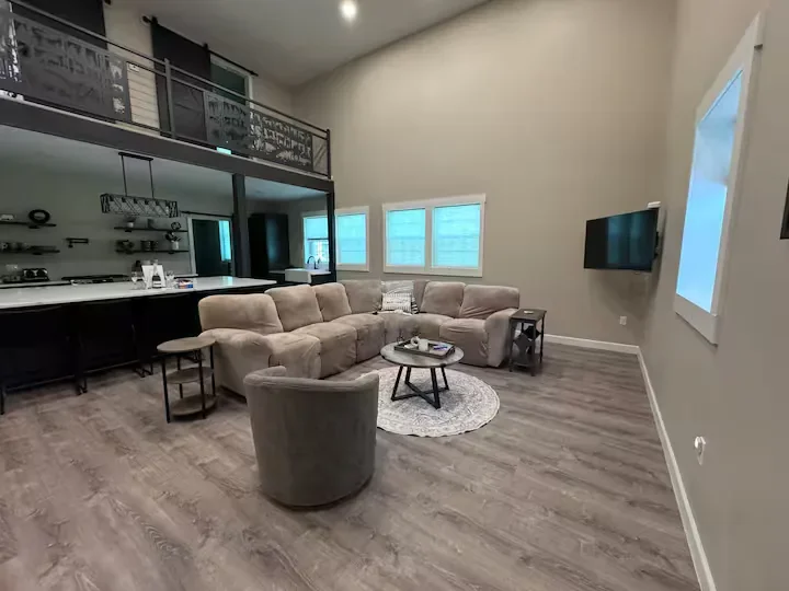 Living room with beige sectional sofa, accent chair, small round coffee table on a rug, wall-mounted TV, and windows. There is a kitchen counter with bar stools in the background.