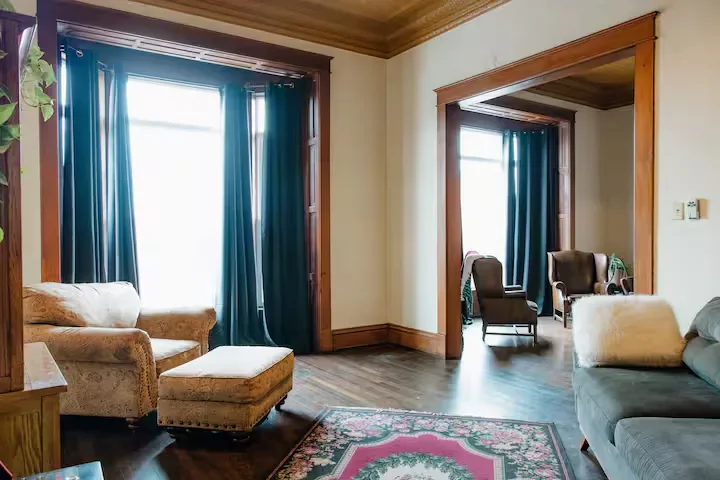 Living room with two large windows with blue curtains, a beige armchair with an ottoman, two dark brown armchairs with green plants on side tables, a patterned area rug, and wooden trim around doorways and window frames.