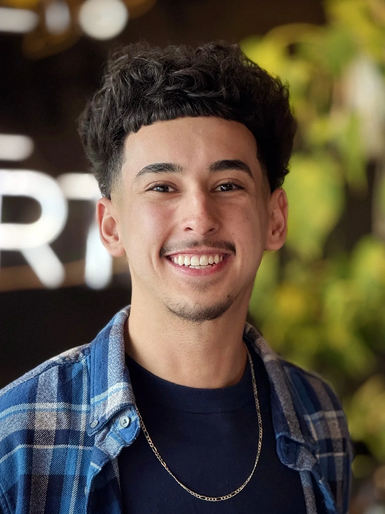 Chris smiling, wearing a black t-shirt and a plaid button down, with a blurred background.