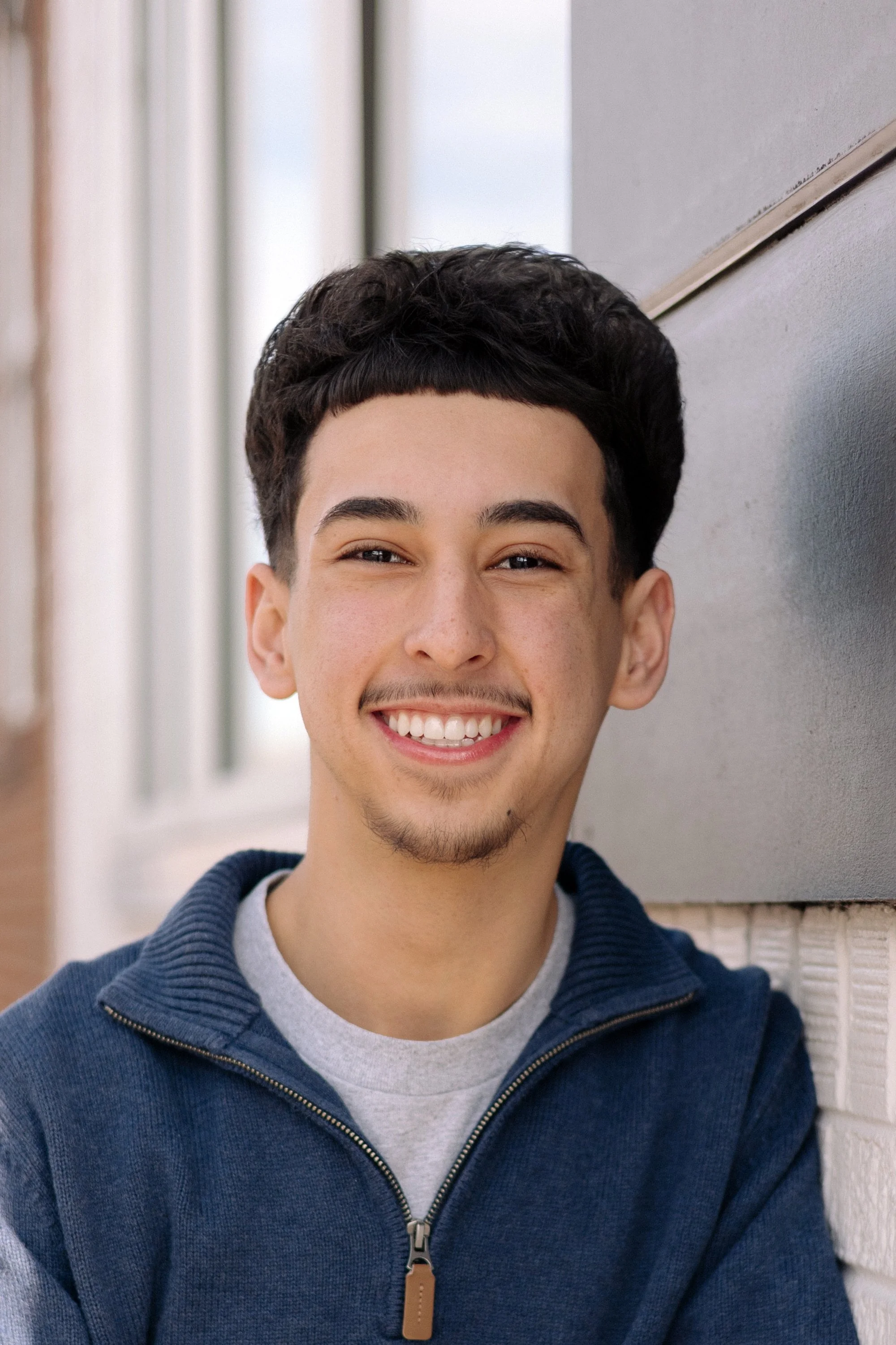 Chris outdoors smiling, wearing a gray t-shirt and a navy sweater, with a blurred gray background.