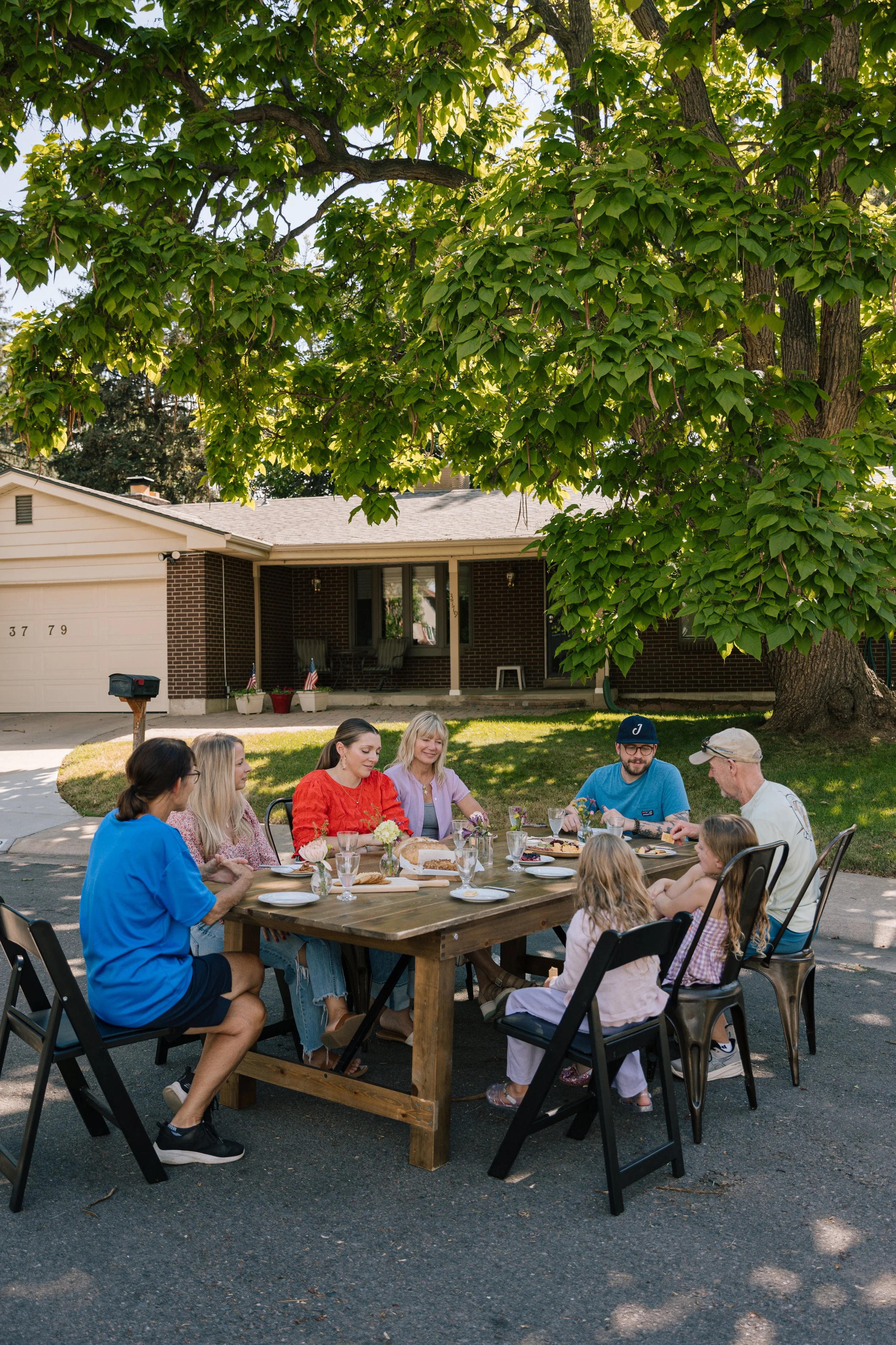 A group of people gathered around a wooden table for an outdoor meal in a suburban neighborhood, with a large leafy tree overhead, and a house with a brick facade in the background.