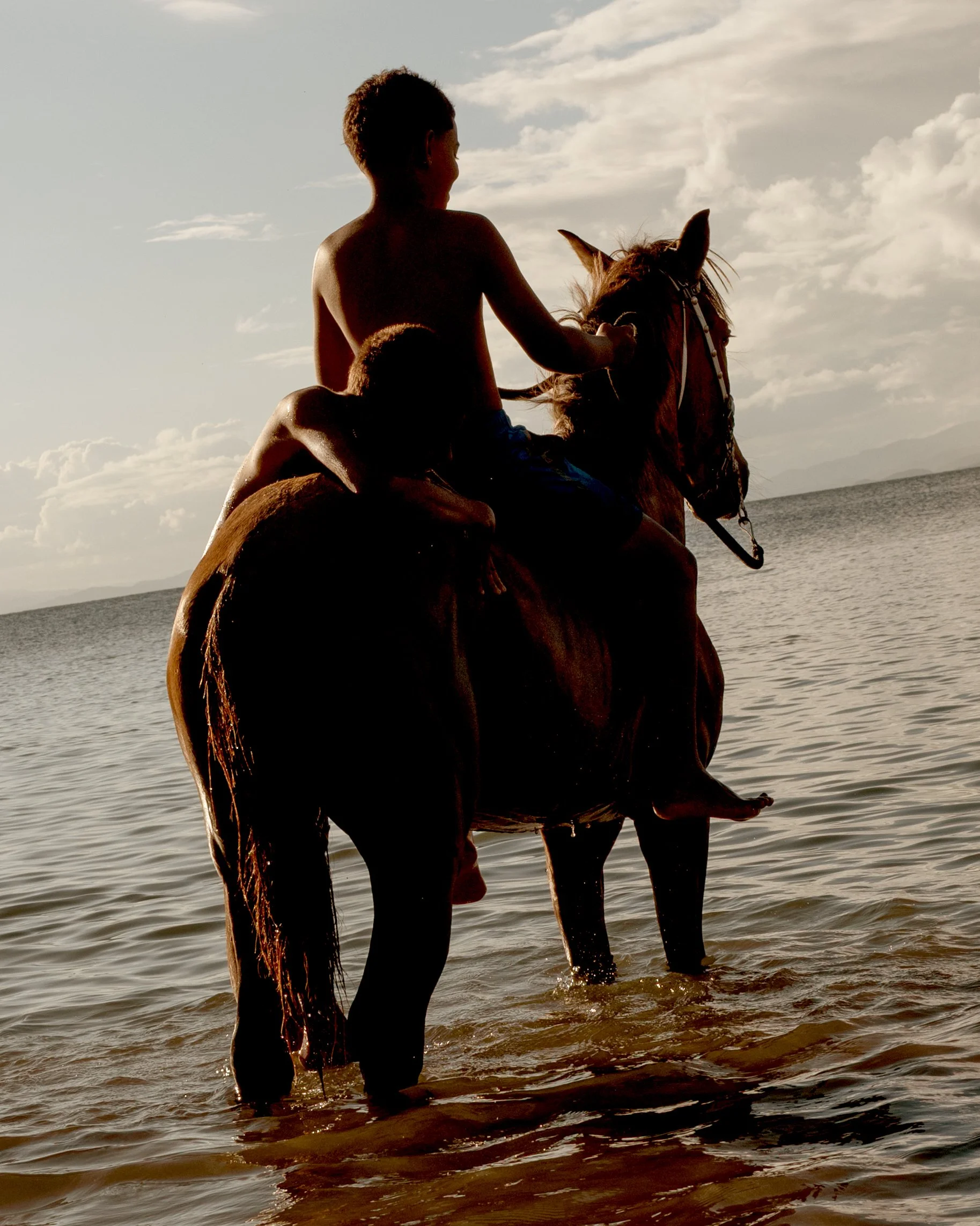 Two children and a horse at the beach, the children riding the horse in the shallow water, with a cloudy sky in the background.