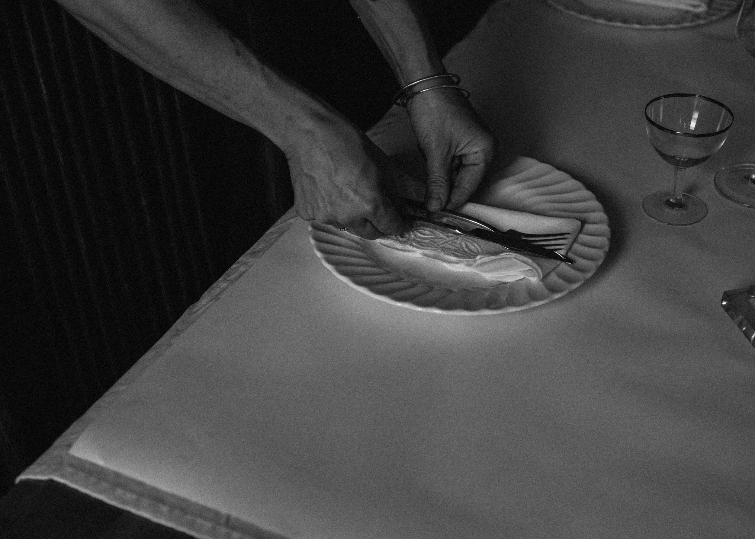 A person setting a table with a plate, fork, knife, and a glass, with a napkin under the utensils, in a dining setting.