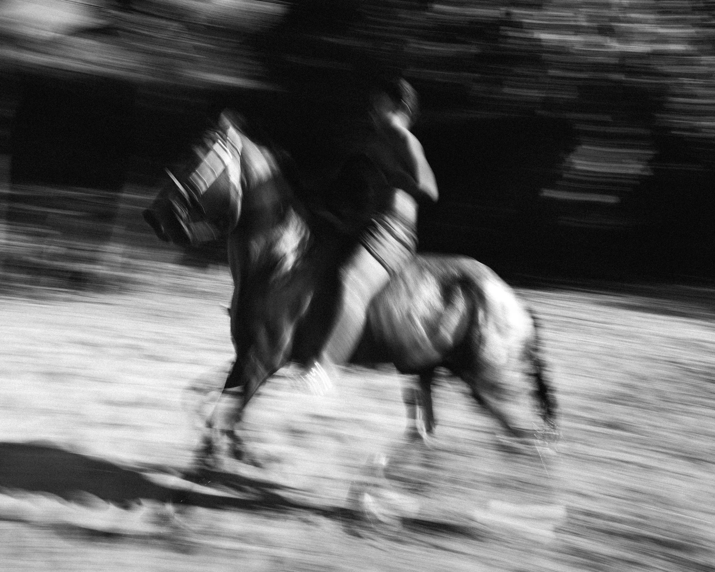 A black and white image showing a person riding a horse at full speed, creating a motion blur effect.