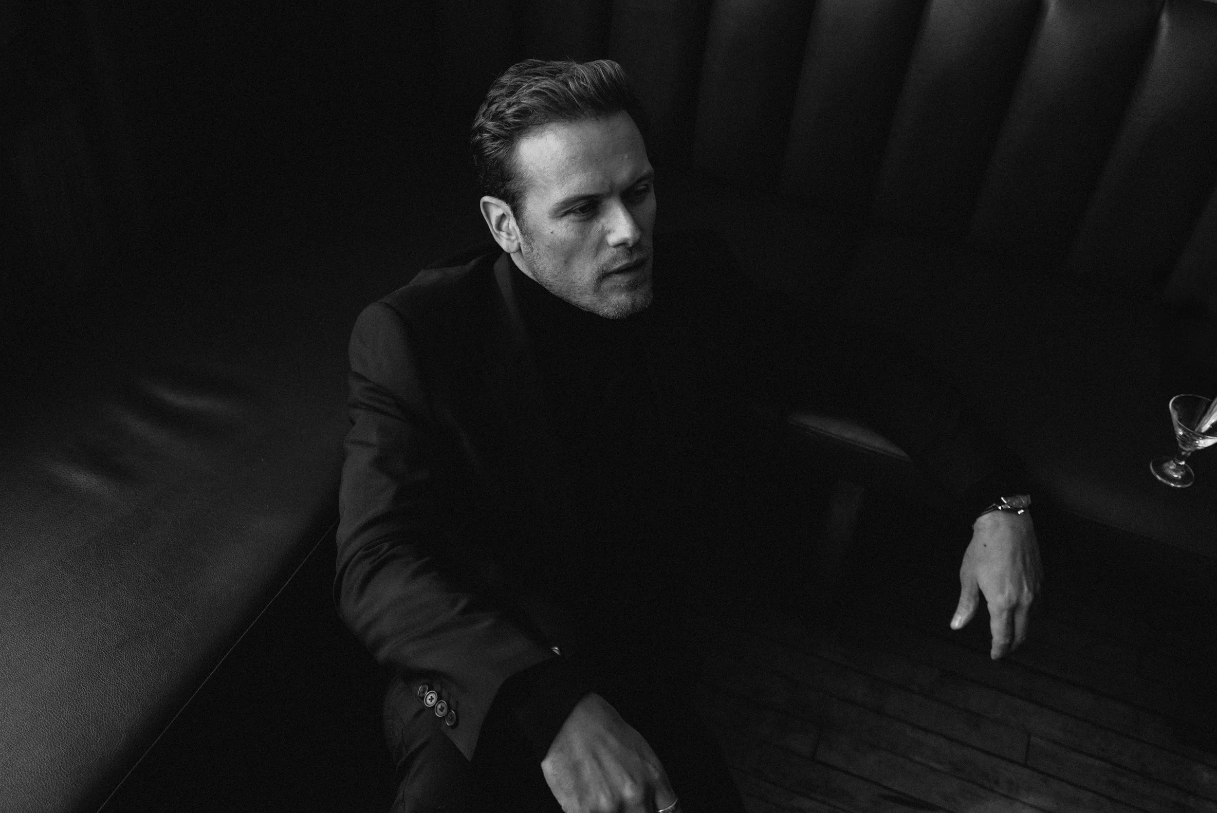 A black and white photo of a man with styled hair and a beard, wearing a dark suit jacket, sitting at a table with a glass of wine and some food. The background is dark with vertical paneling.