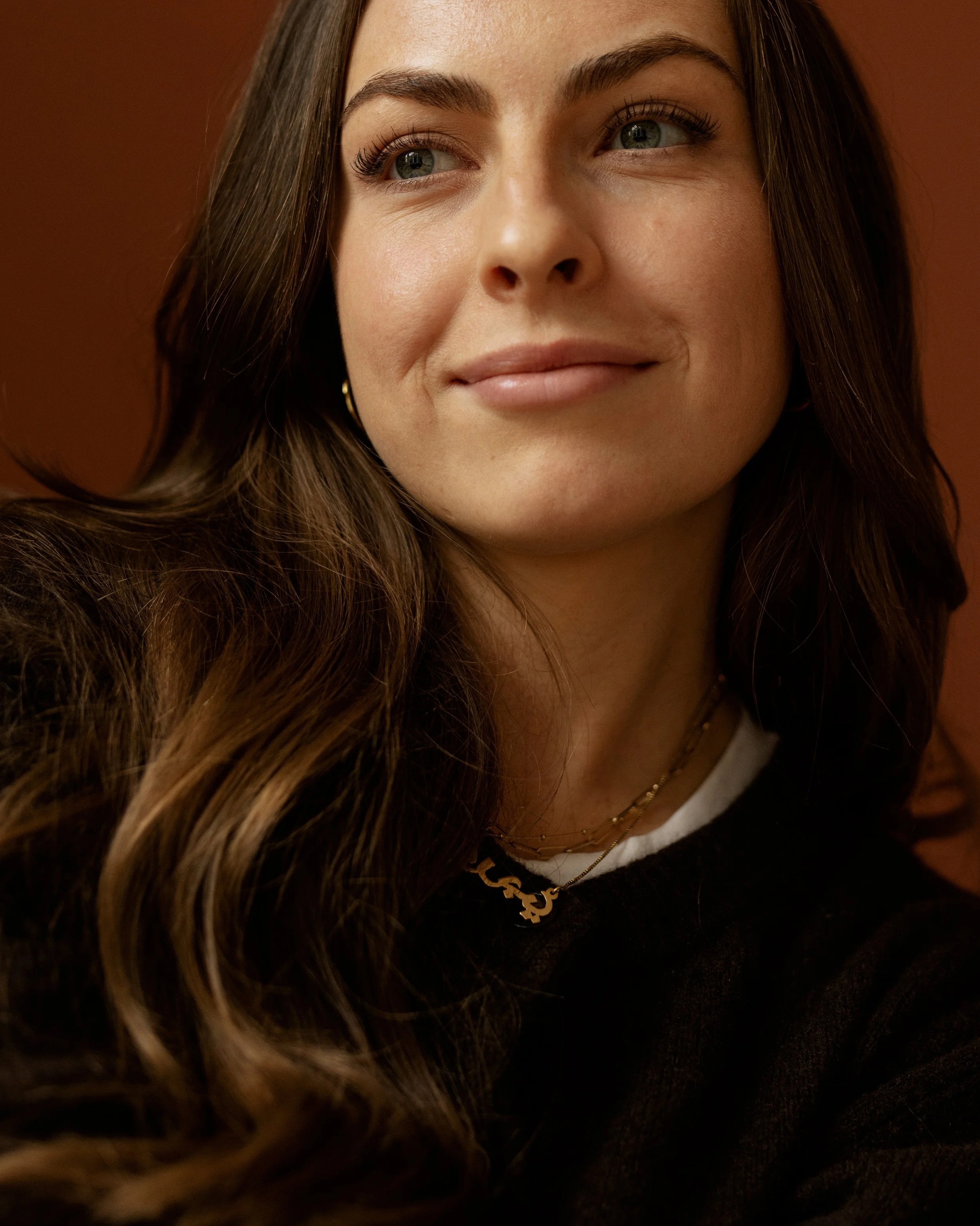 Close-up of a young woman with long wavy hair, wearing gold earrings and layered necklaces, smiling softly, with a warm background.