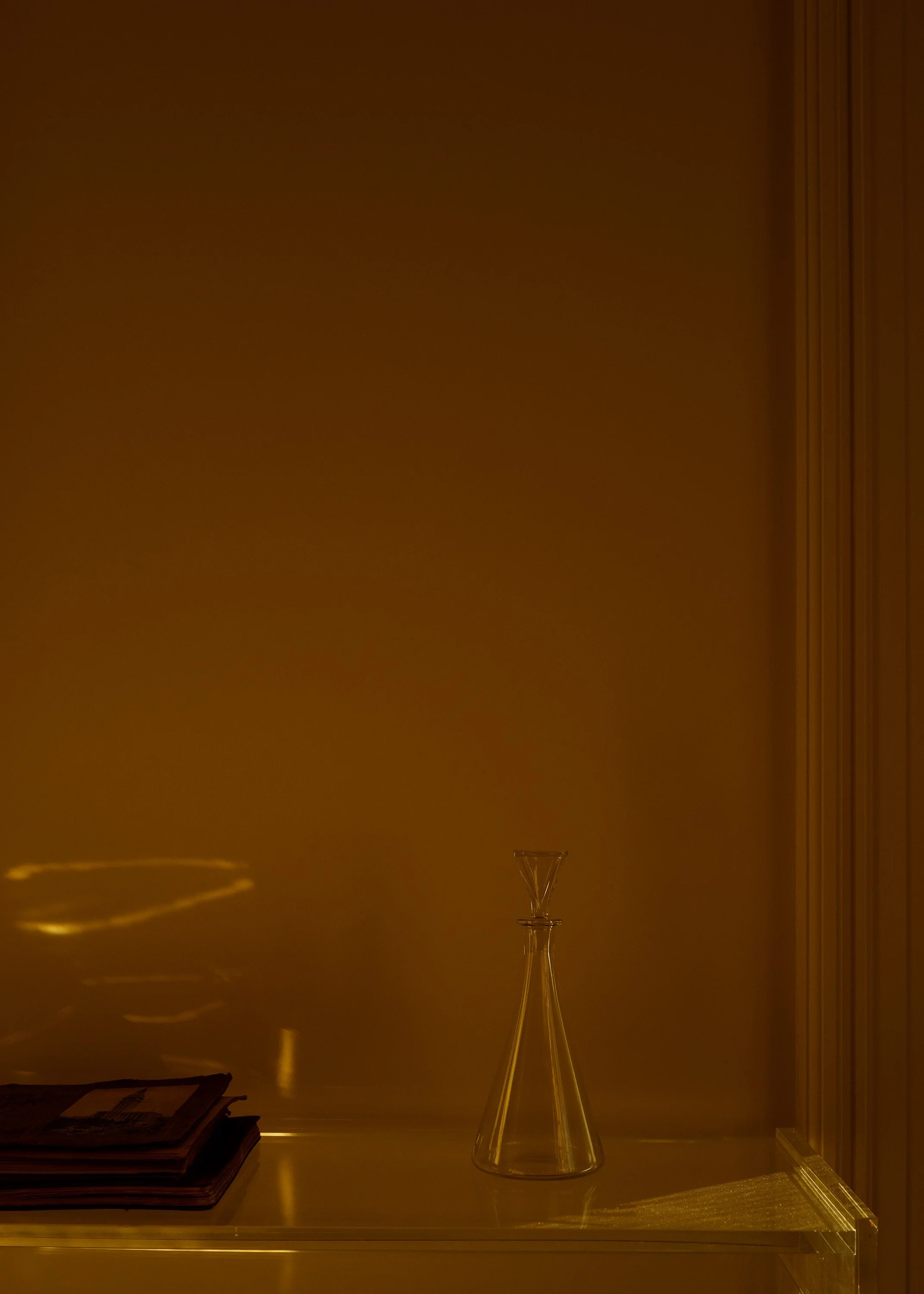 A clear glass champagne flute on a table with a reflection, set against a plain wall with vertical blinds and a small pile of magazines or books nearby.