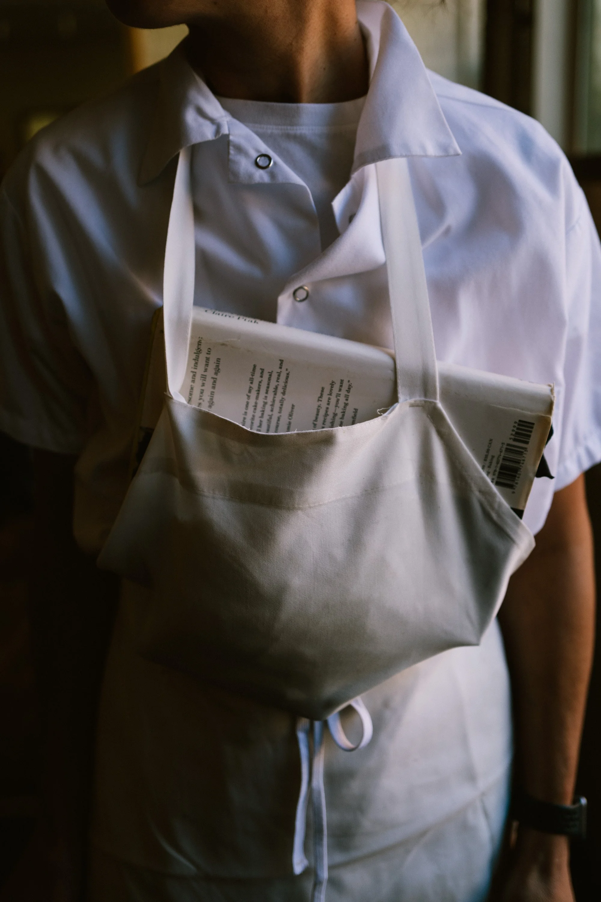 A chef wearing a white button-up shirt and gray chef pants, holding a book with text on the page, with part of their face and arm visible.