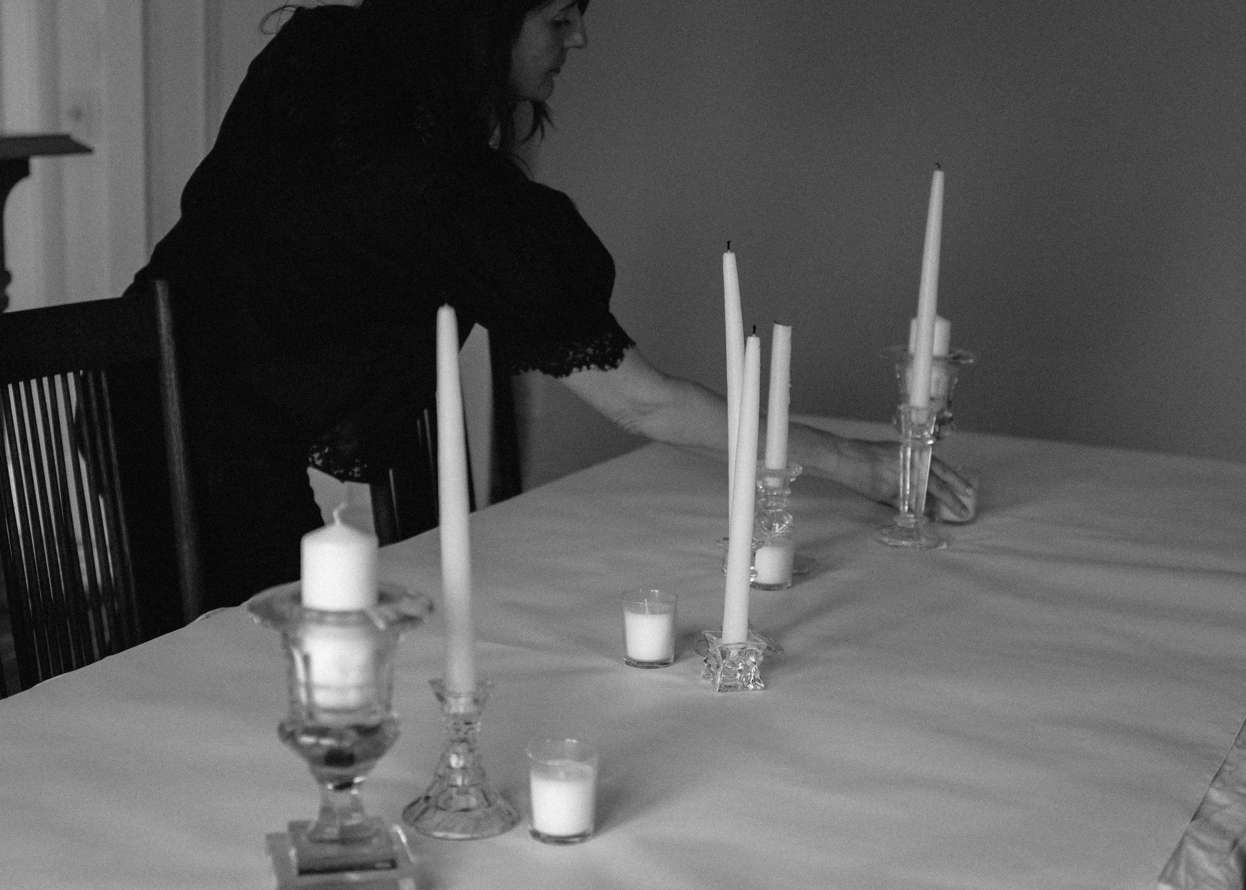 A woman with dark hair sitting at a dining table with tall white candles in glass candleholders, in a dimly lit room.