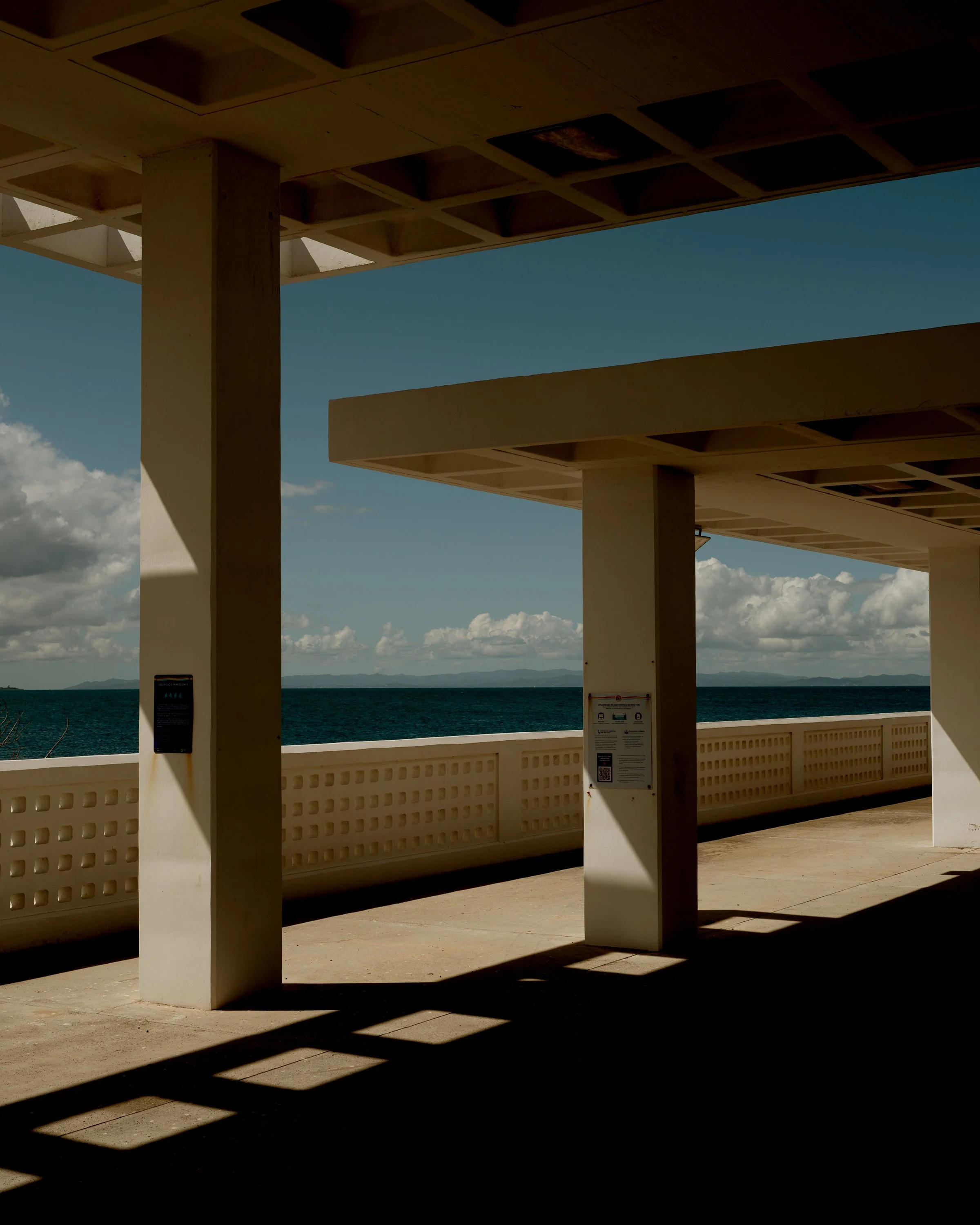 A covered outdoor structure overlooking the ocean with a blue sky and clouds in the background. The structure has concrete columns and a ceiling with rectangular openings, casting shadows on the ground and a decorative wall with square cutouts.
