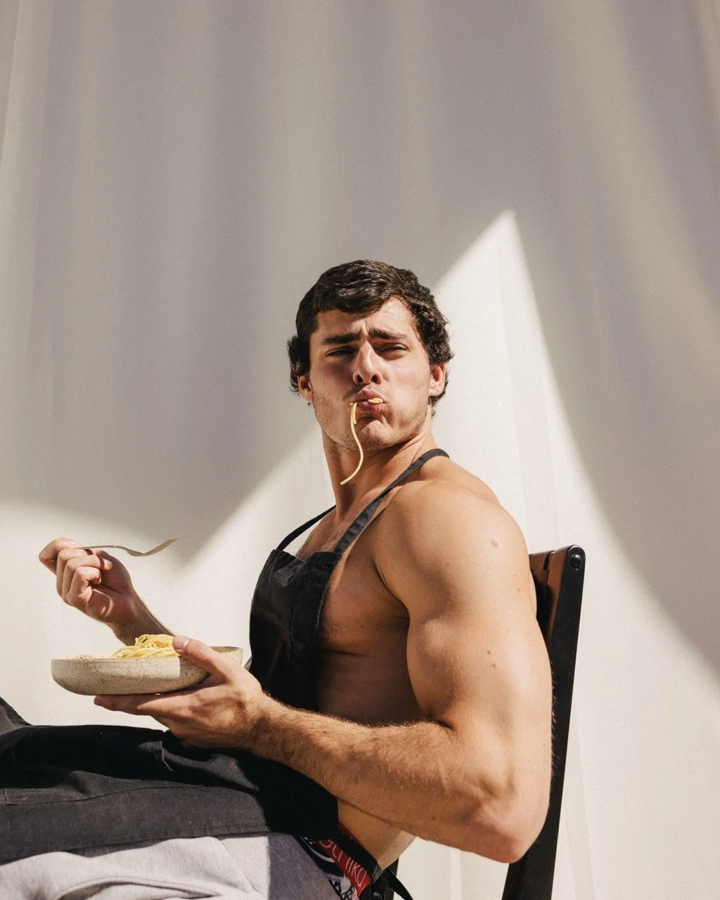 A shirtless young man with dark curly hair wearing an apron, sitting on a dining chair, eating spaghetti with a fork in one hand and holding a bowl of pasta in the other, looking at the camera with a playful expression.