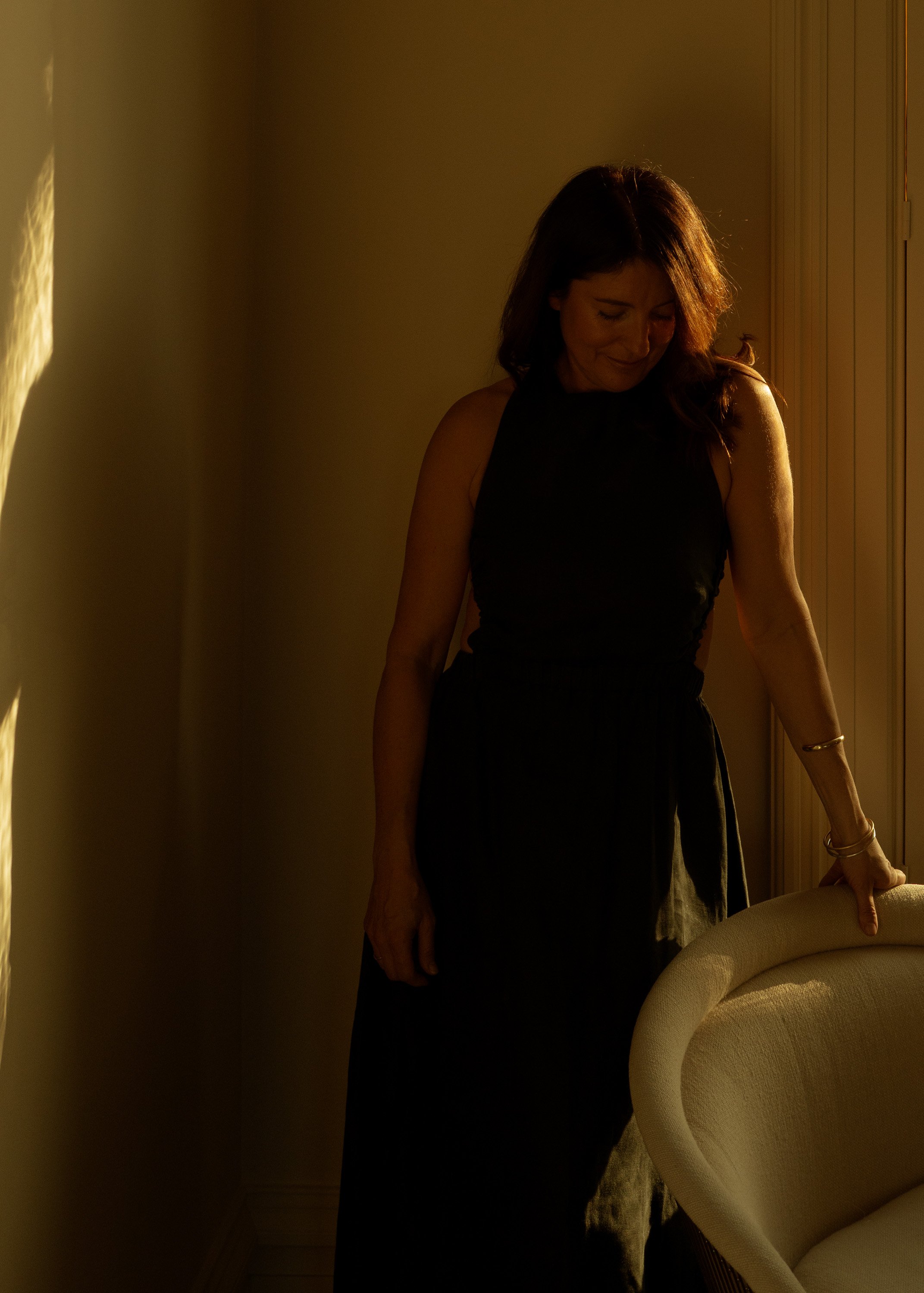 A woman with dark brown hair wearing a black dress standing in a dimly lit room, smiling with her eyes closed, holding the back of a beige chair.