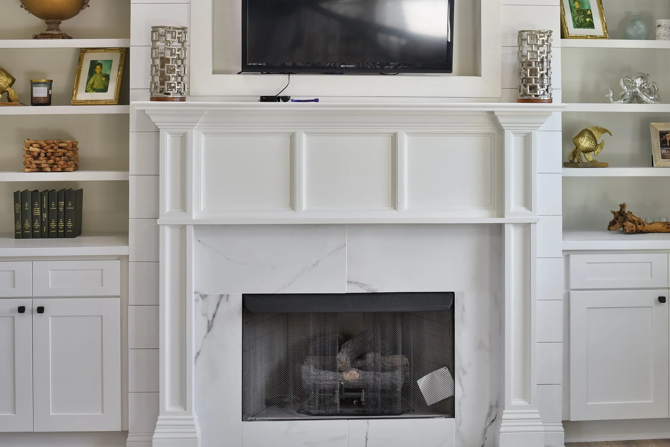 A white fireplace with a marble finish, flanked by built-in shelves with various decorative items, and a flat-screen TV mounted above