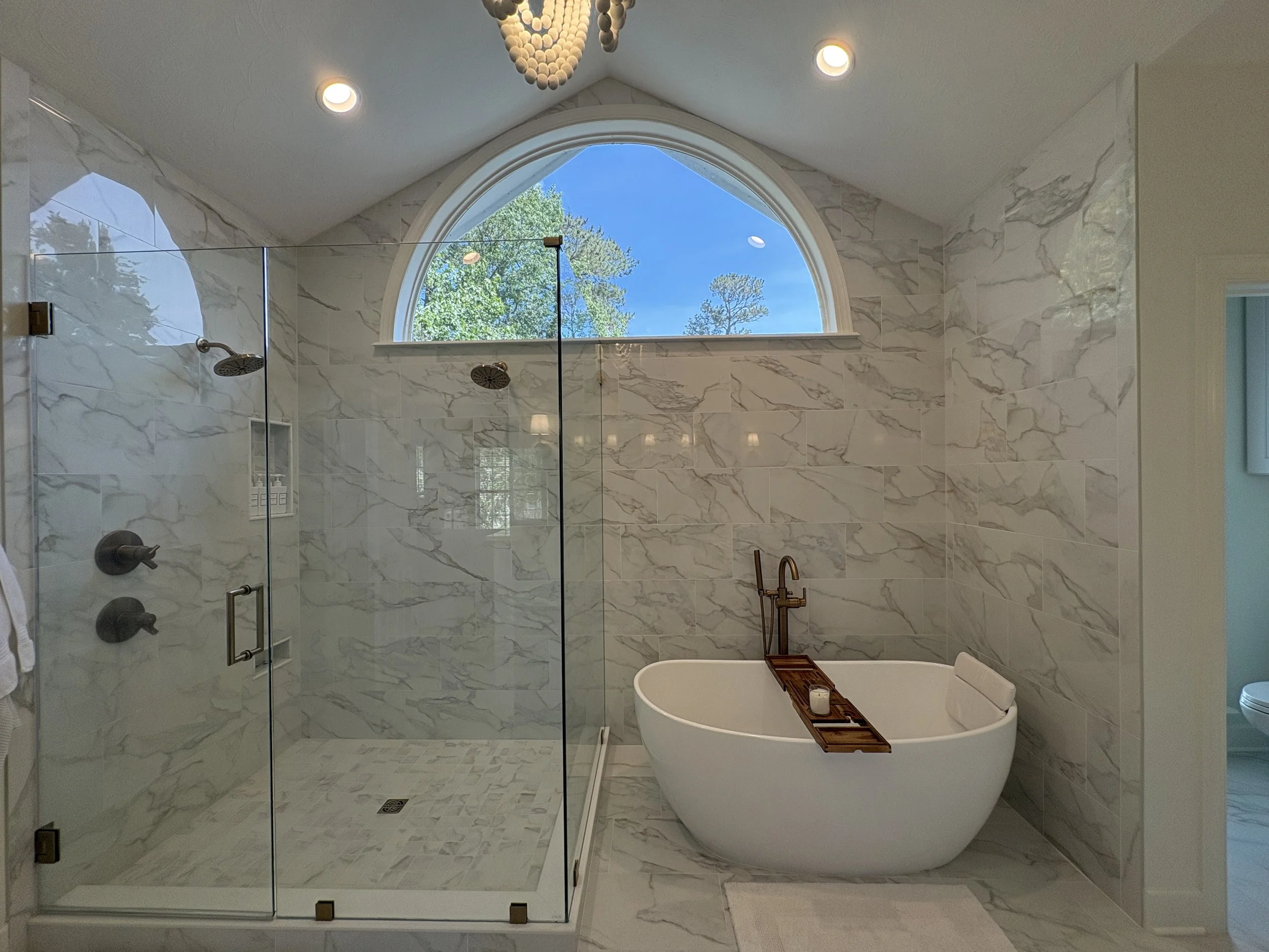 Finished primary bathroom featuring a freestanding soaking tub, frameless glass walk-in shower, and floor-to-ceiling marble-look tile that creates a bright, open, and luxurious atmosphere. An oversized arched window fills the space with natural light