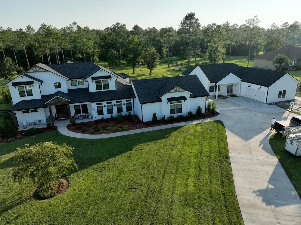 A large white modern house with a black roof, surrounded by a spacious green lawn and trees, with a long driveway leading to a garage and a person near the driveway.