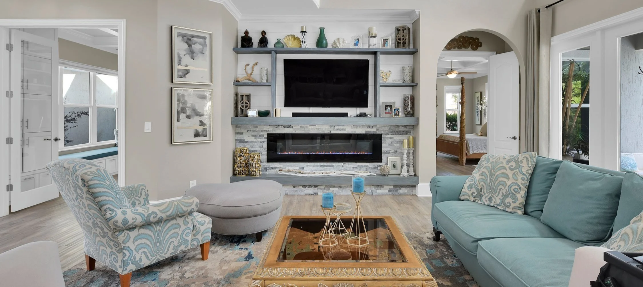 Living room with a blue sofa, patterned armchair, glass-top coffee table, and a modern fireplace with a TV mounted above. Decorative items on shelves and windows with curtains in the background.