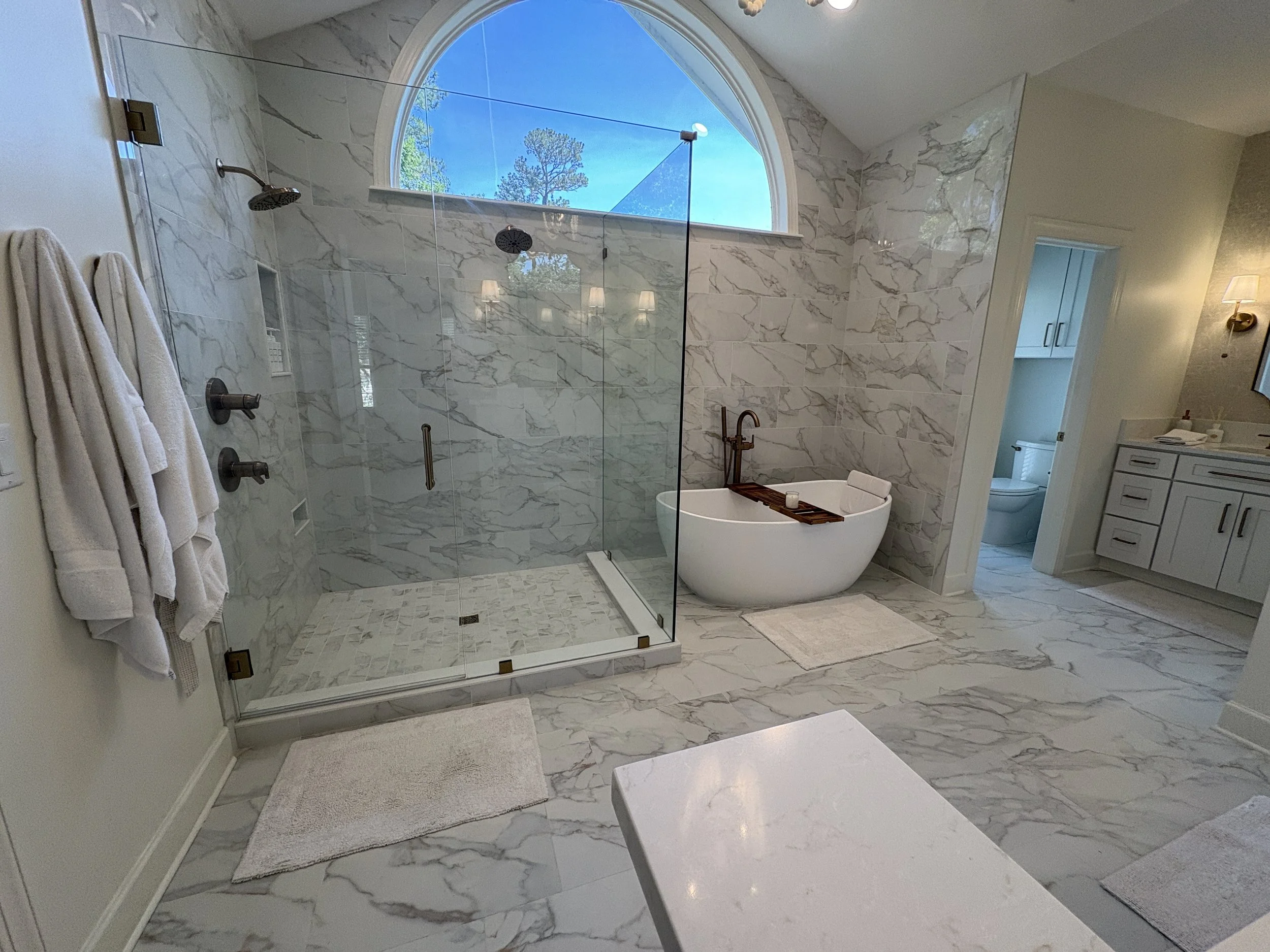 Finished luxury primary bathroom showcasing a spacious frameless glass walk-in shower, freestanding soaking tub, and floor-to-ceiling marble-look tile throughout the floors and walls. The open-concept layout, oversized arched window, and premium bron