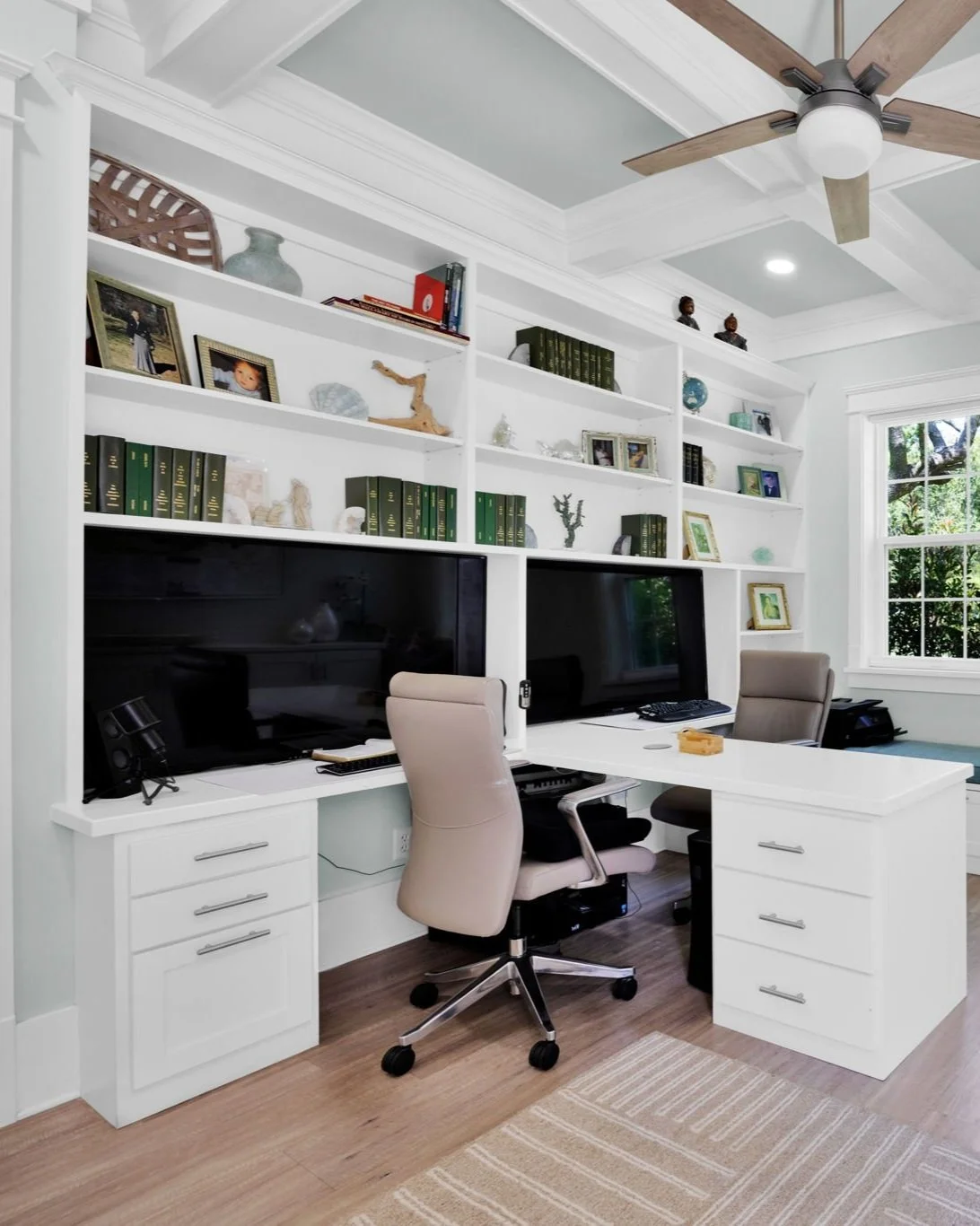 Home office with white built-in desk and shelving, two large computer monitors, beige office chairs, and decorative items including photo frames and small sculptures. Natural light from a window illuminates the space, with a ceiling fan above and woo