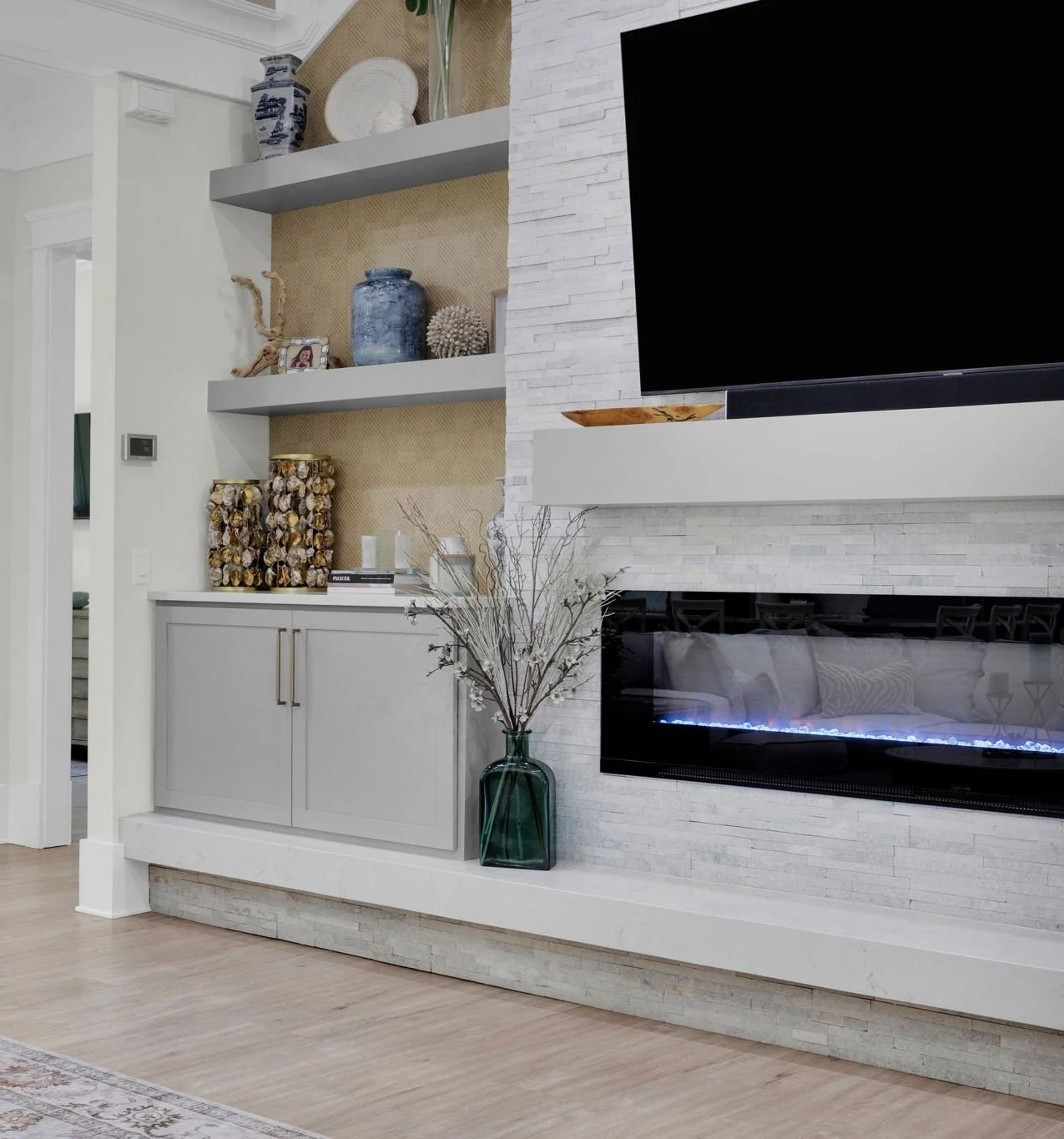 Living room with mounted TV above a modern fireplace, decorative vases and objects on open shelves, and a large glass vase with dried branches on the floor.