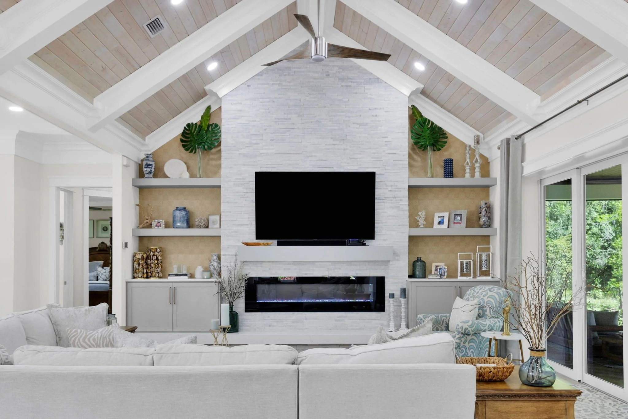 Living room with vaulted ceiling, ceiling fan, white sofa, built-in shelves with decorative items, fireplace, TV, and large sliding glass doors showing greenery outside.