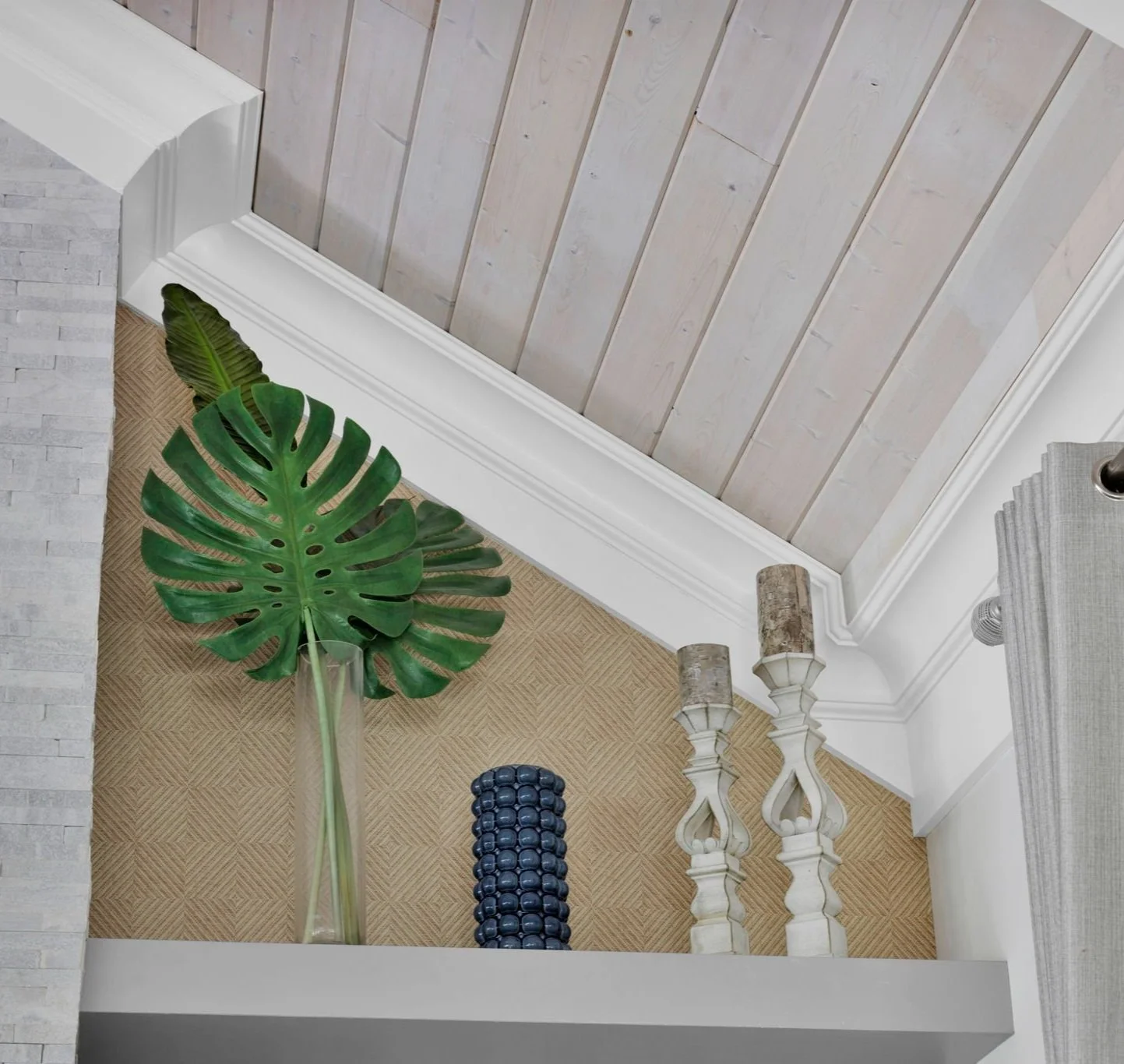 Decorative shelf with large green tropical leaf in a tall glass vase, a blue beaded decorative object, and two white candlesticks with candle holders against a background of light textured walls and a beige patterned rug.