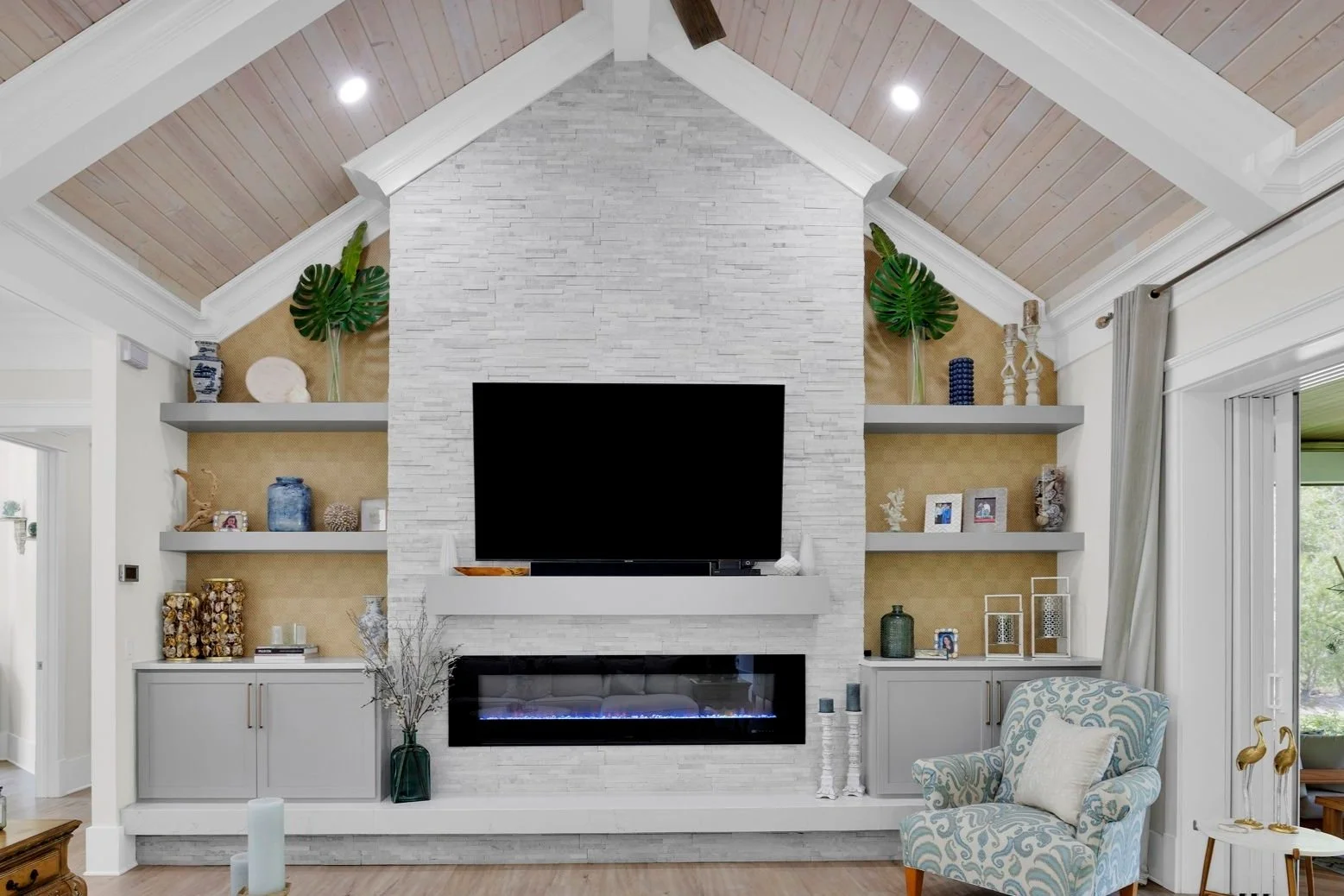 Living room with white brick fireplace, mounted flat-screen TV, built-in shelves with decorations, gray cabinets, light wood vaulted ceiling, armchair, and curtains.
