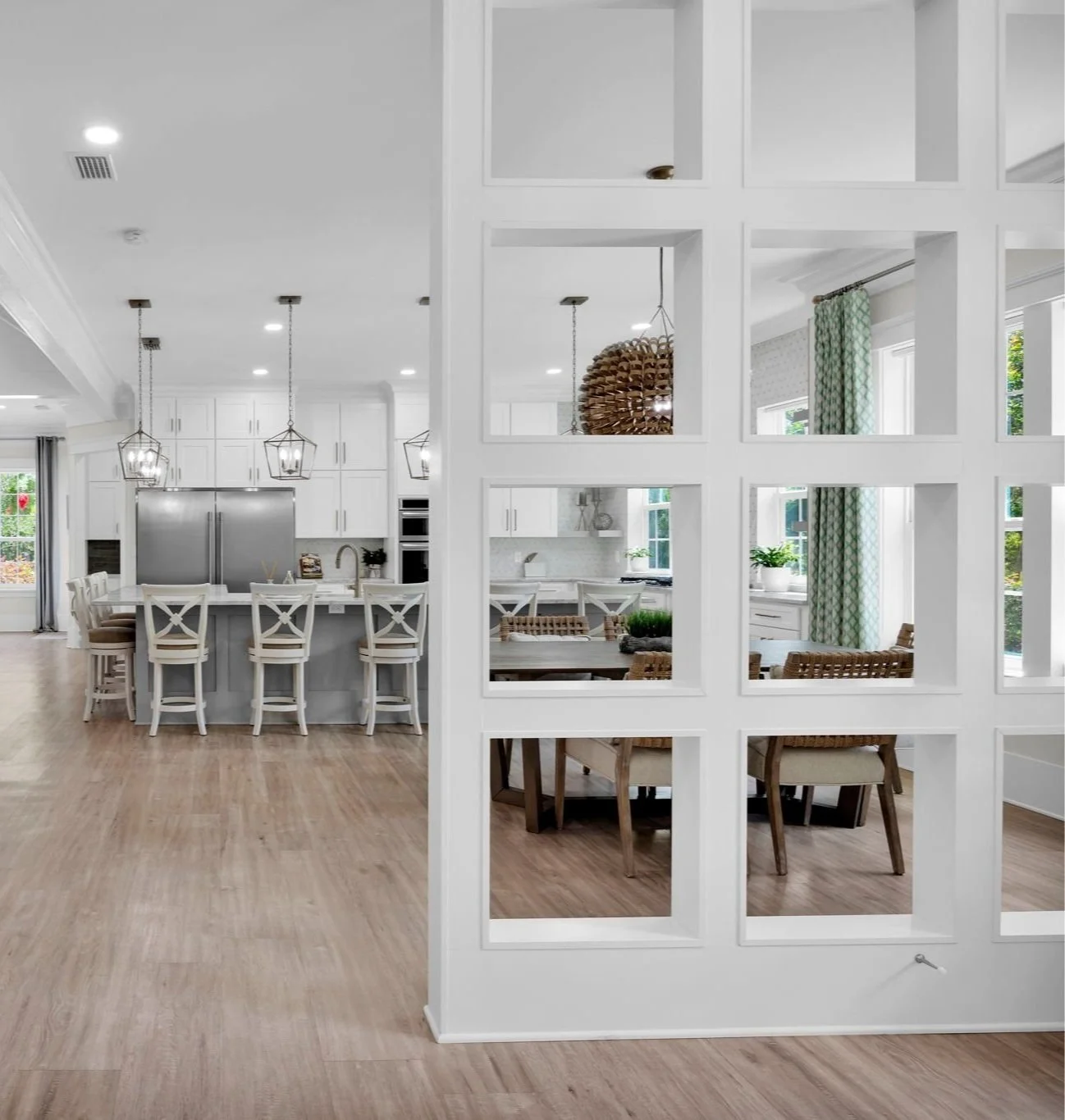 Open-concept kitchen and dining area with white cabinetry, stainless steel refrigerator, island with barstools, wooden dining table with chairs, and large windows with green curtains.