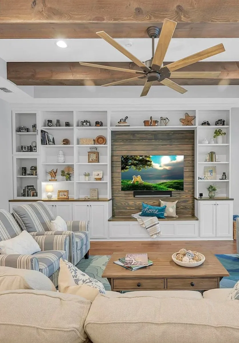 Living room with built-in white shelves, a flatscreen TV, a beige sofa, and a wooden coffee table. The TV is surrounded by wooden paneling, and the ceiling features a large wooden beam with a ceiling fan. Decor includes books, cameras, vases, and dec