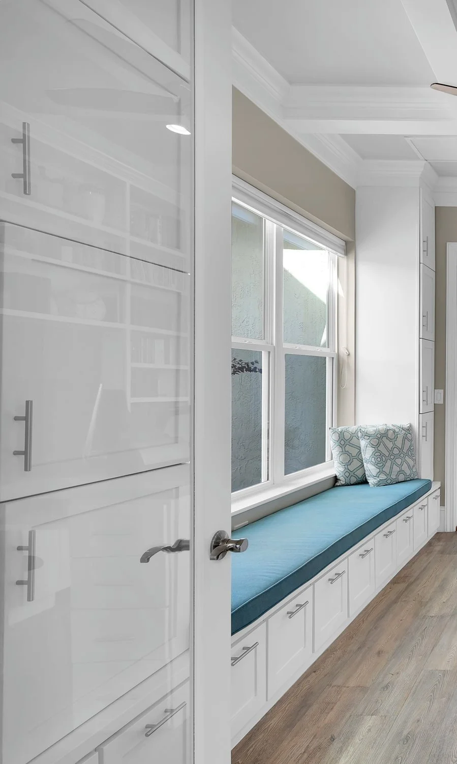 Sunlit window seat with blue cushion and patterned pillows, built into a white wall with cabinetry below, in a bright room with hardwood floors.