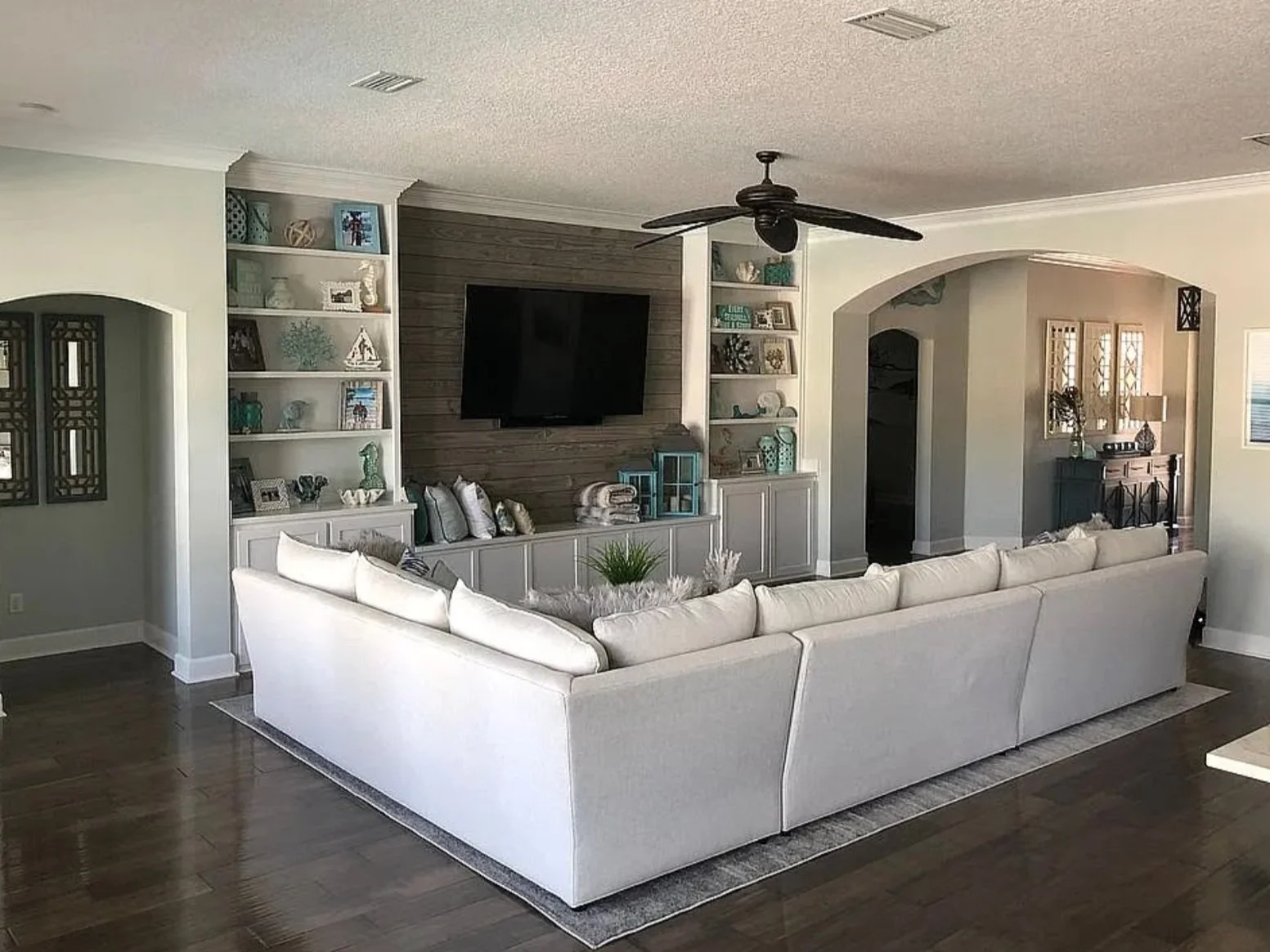 Bright living room with white sectional sofa, wooden floor, built-in shelves with decor, large TV on wall, ceiling fan, and a hallway leading to other rooms.