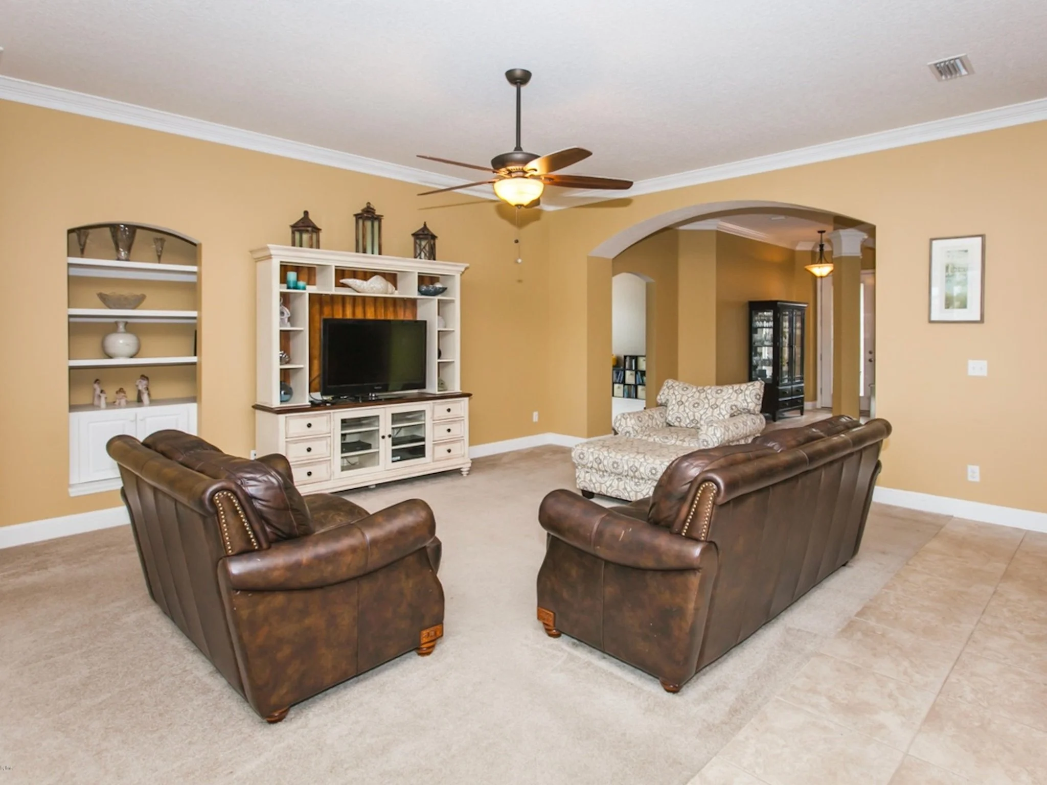 Living room with two brown leather sofas, a patterned armchair, a white entertainment center with a flat-screen TV, built-in shelves, and decorative lanterns on top. Beige walls with white crown molding and a ceiling fan.