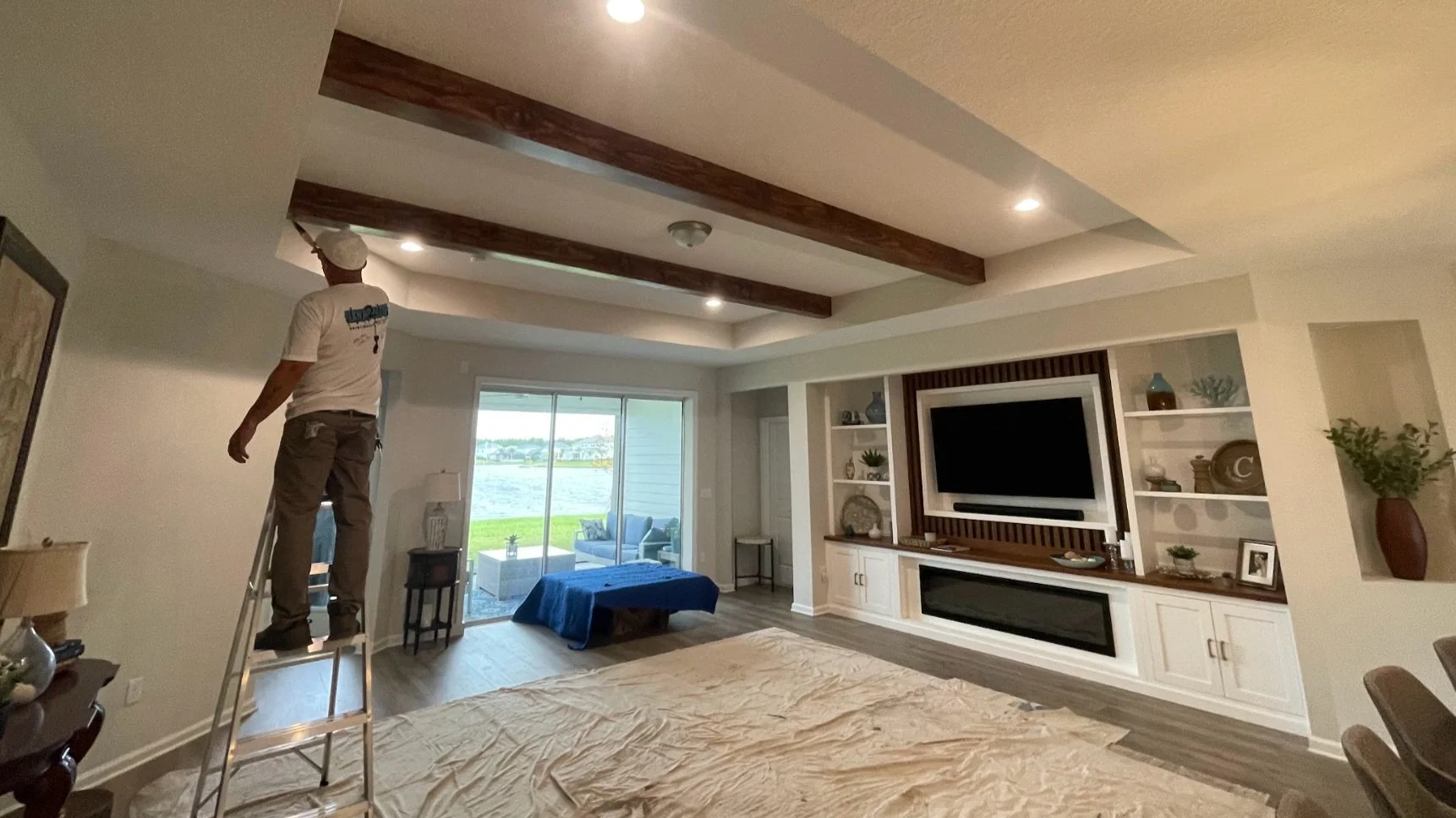 A man standing on a ladder working on the ceiling of a living room with a large TV, sliding glass door, and outdoor patio area visible.