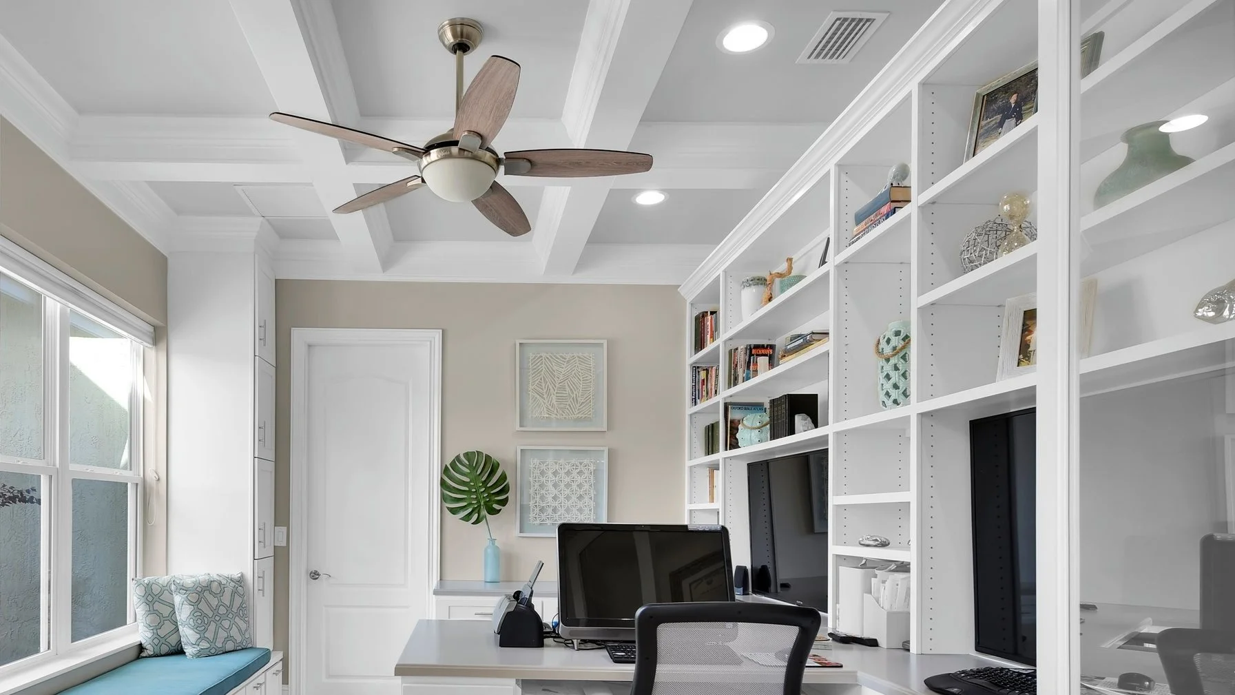 Home office with white built-in shelves, desk with a computer and office supplies, a ceiling fan, a large window with a window seat, beige walls, and decorative items on the shelves.