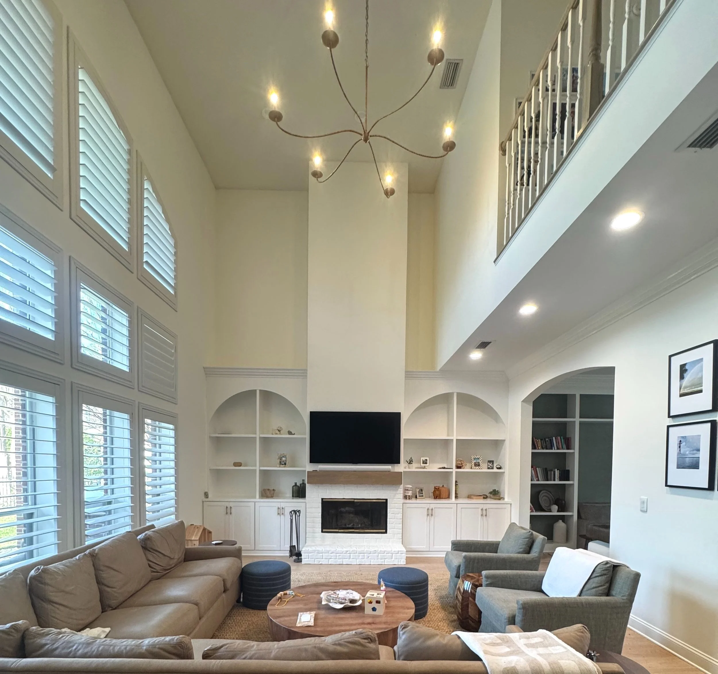 Living room with a high ceiling, large windows with white shutters, a beige sectional sofa, a wooden coffee table, two gray armchairs, a fireplace with a mounted TV, built-in white shelves, and a staircase with a balcony railing.