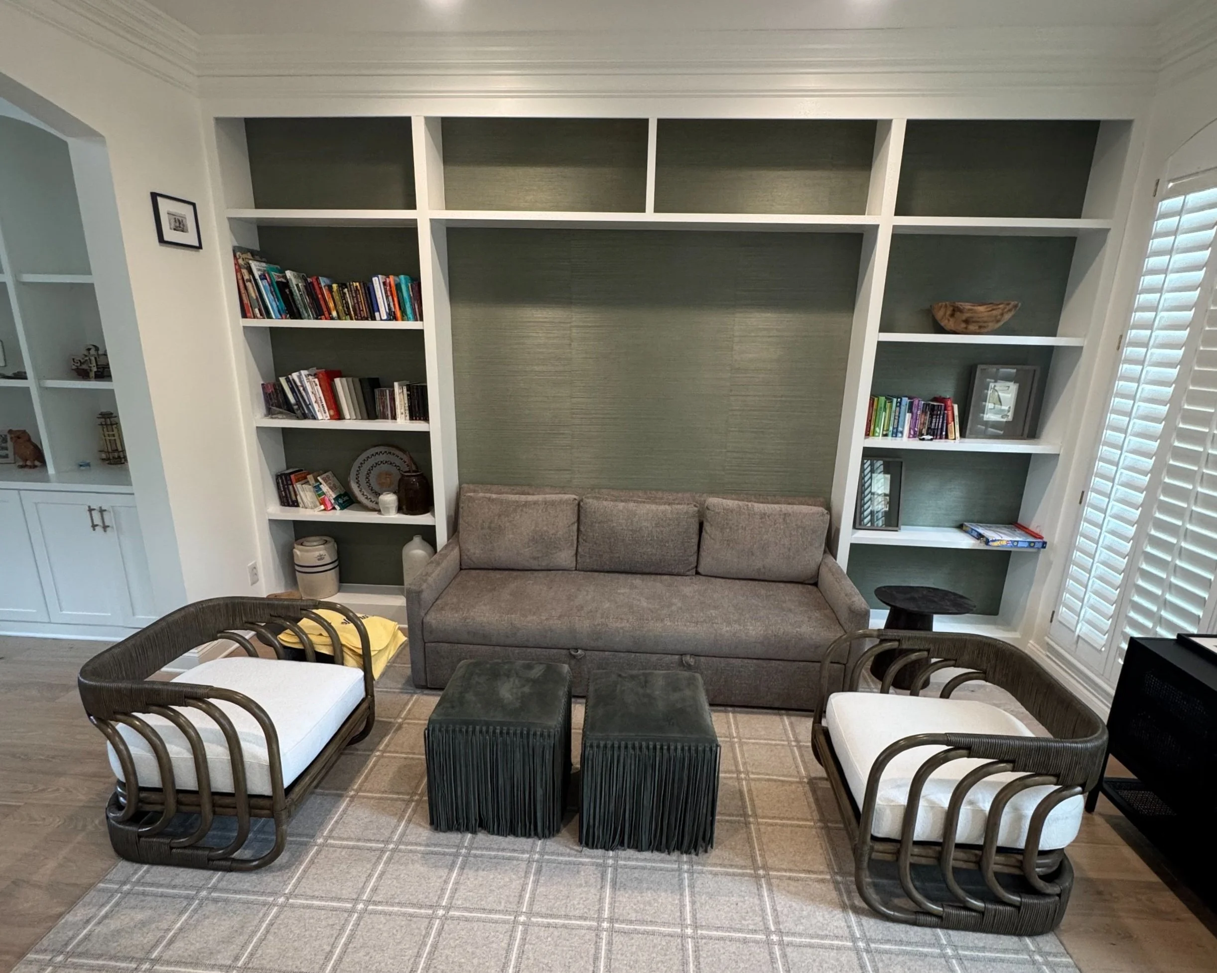 Living room with built-in bookshelves, a gray sofa, two armchairs with white cushions, and two green ottomans. The room has a patterned rug and a window with white shutters.