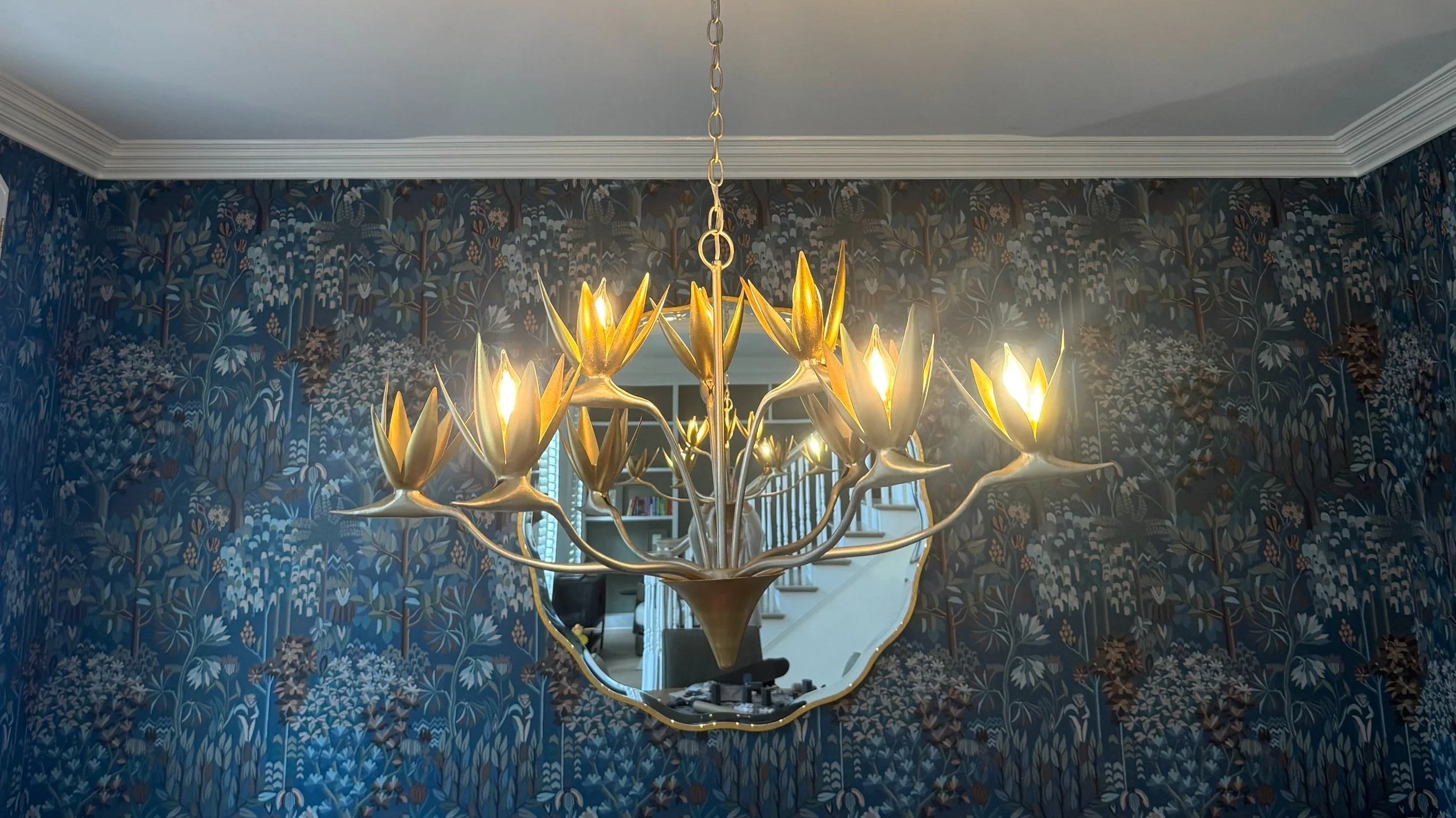 A chandelier with a floral design hanging from a ceiling in front of a wallpapered wall with a botanical pattern.