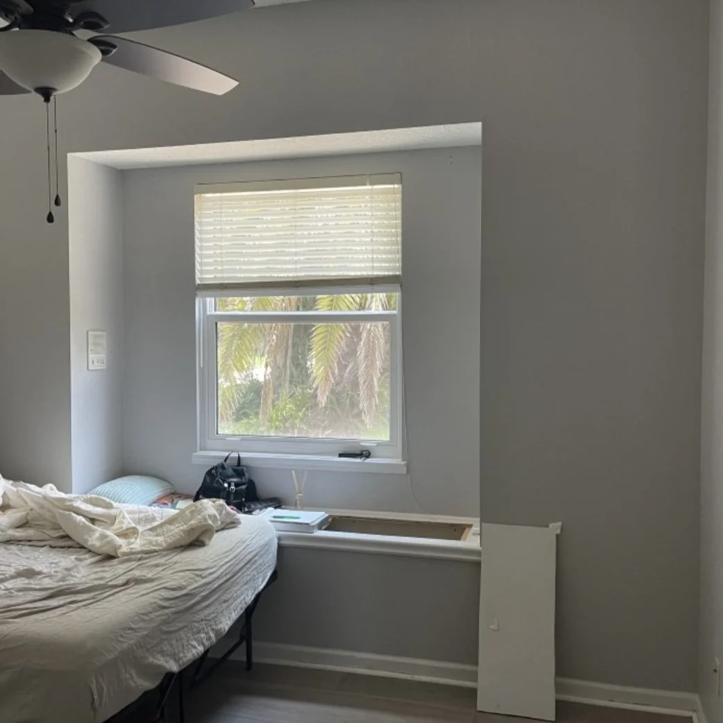 A bedroom with a bed, window, and a ceiling fan. The bed is unmade, and there are some items on the window sill, including a black bag and a laptop. The window has blinds, and outside there are trees with green leaves.