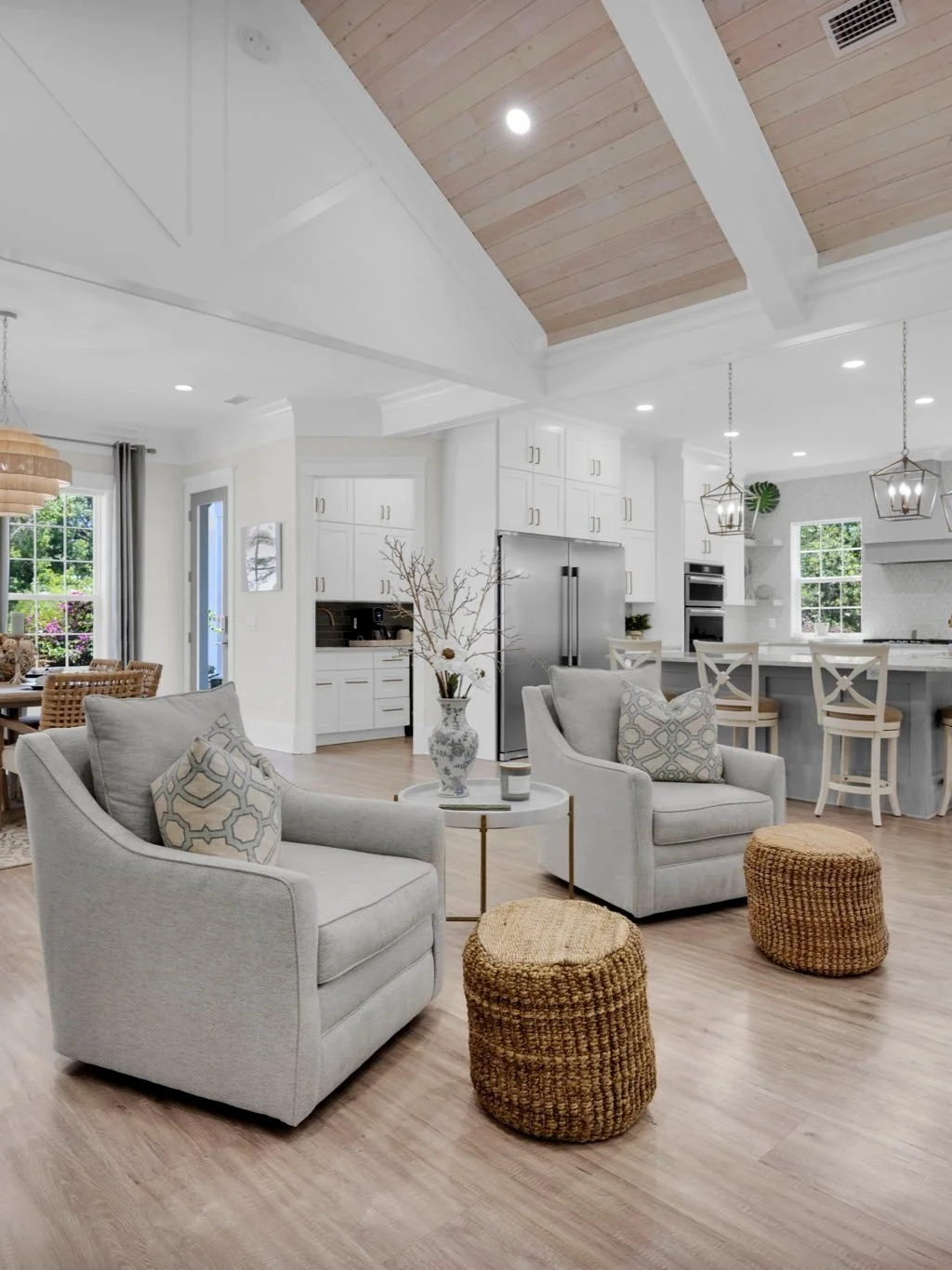 Bright open-concept living space with white walls, light wood flooring, and high vaulted ceiling with wood panels; includes gray upholstered armchairs, woven poufs, a small side table with decorative items, and views of the dining area and kitchen wi