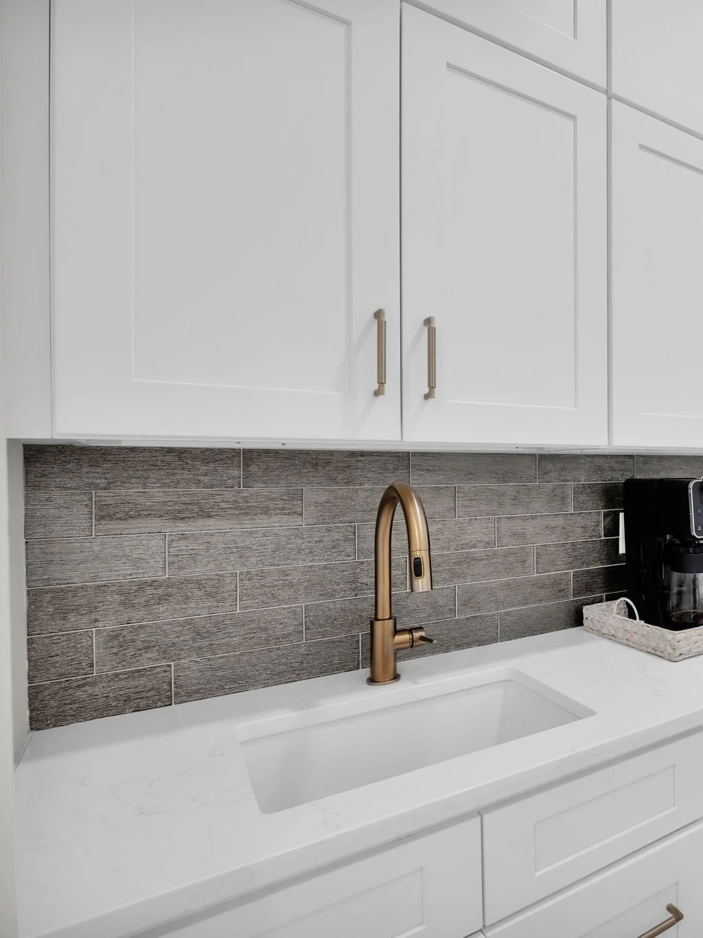 Modern kitchen sink with a brass faucet, white countertop, white cabinetry, gray tiled backsplash, and a coffee maker on the right.