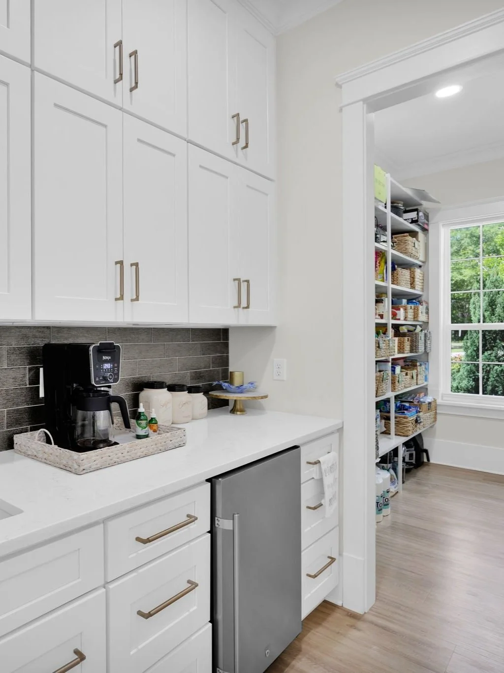 Kitchen with white cabinetry, black backsplash, coffee maker, jars, and a medical air pump, with a door leading to a pantry filled with baskets and supplies.