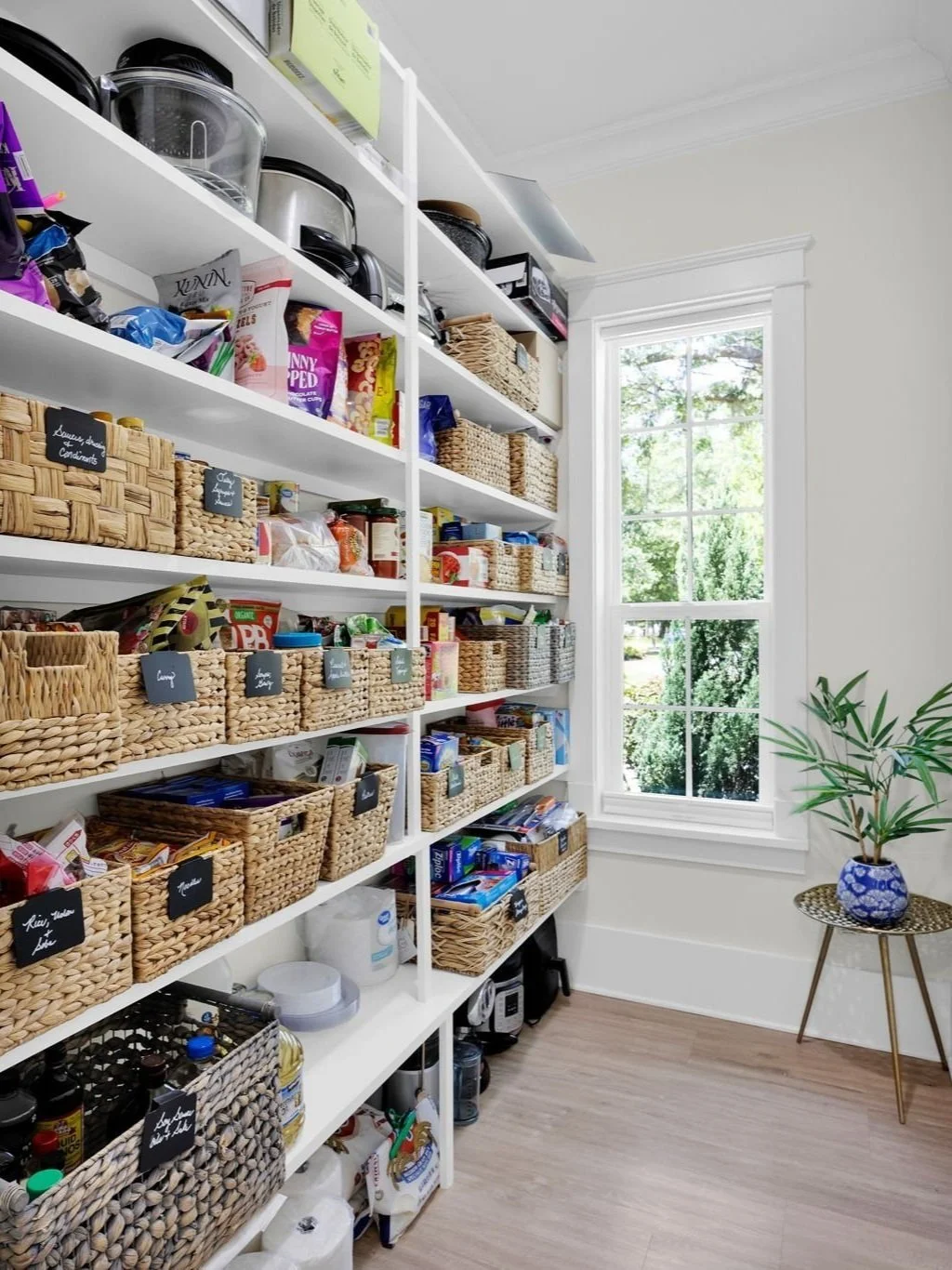 Pantry with white shelves filled with woven baskets containing snacks and canned goods, next to a window with greenery outside and a potted plant on a small table.