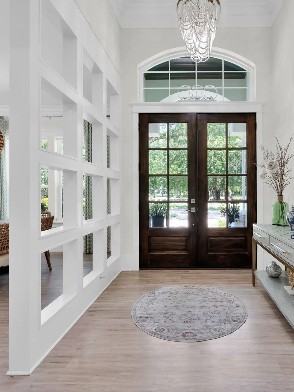 Entryway of a house with dark wooden double doors, large glass windows, a chandelier hanging from the ceiling, a decorative rug on light wood flooring, and a side table with vases and floral arrangements.
