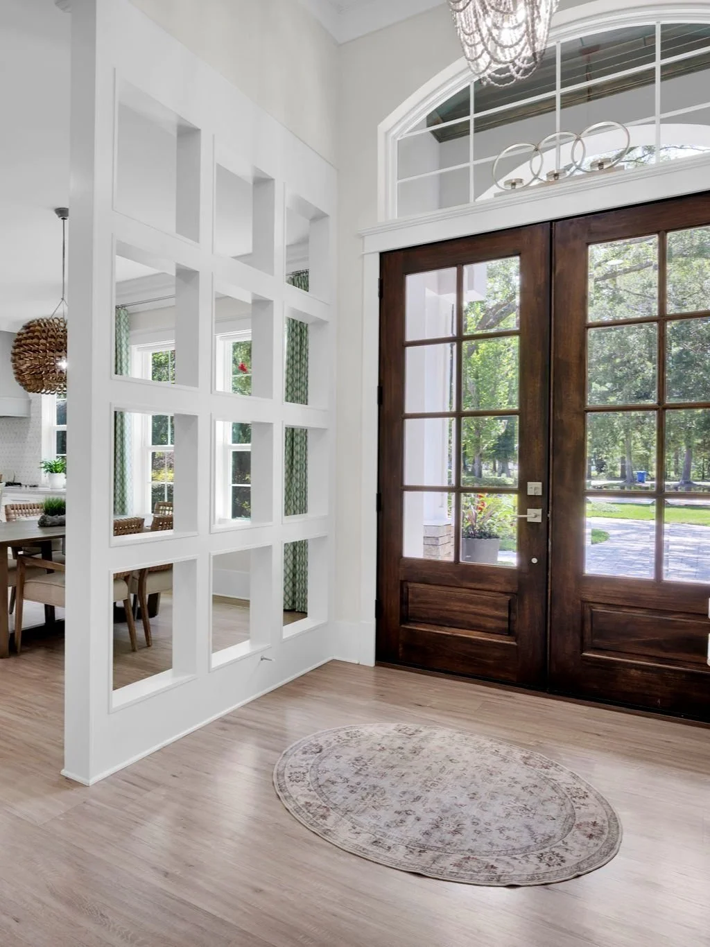 Entrance with dark wooden double doors with glass panes, leading outside to a lush green yard. Bright interior with white walls, light wood flooring, and a small round rug near the door. A decorative chandelier hangs from the ceiling, and a window wi