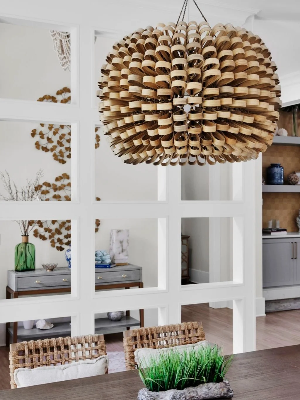 A large, spherical wooden chandelier hanging from the ceiling in a modern dining room, with a side view of a glass with a plant and a decorative green bottle on a surface.