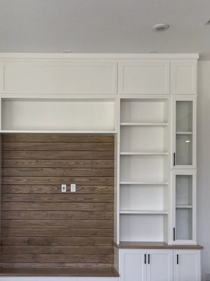Built-in white storage with open shelves and cabinets, featuring a wooden accent wall with electrical outlets.