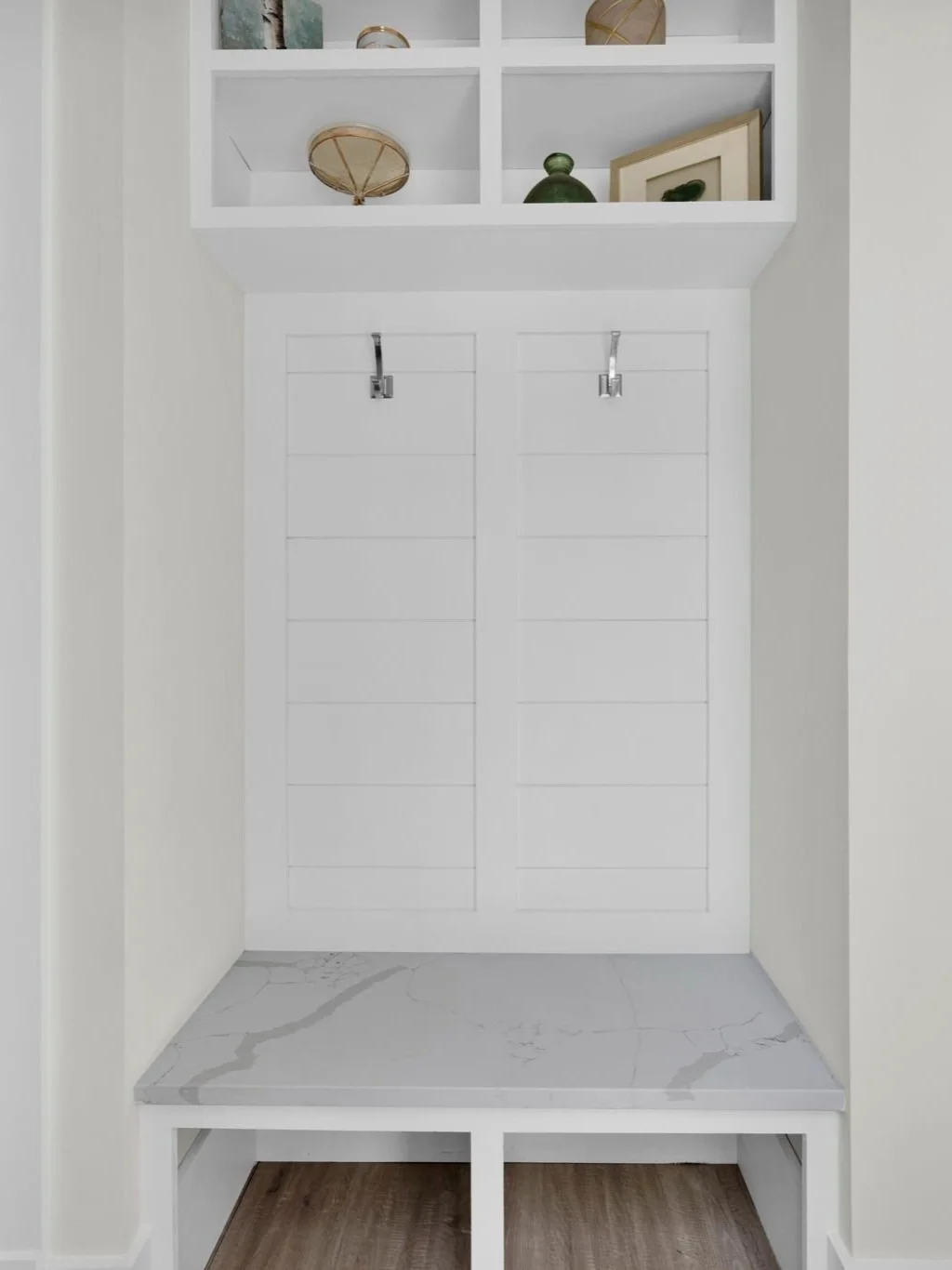 A white built-in entryway bench with a marble top, situated between two white walls, with upper open shelving displaying decorative items.