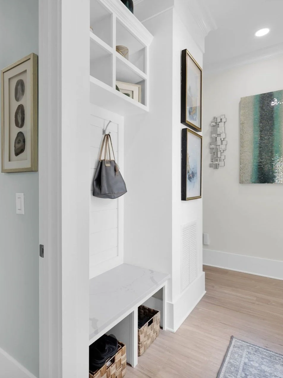 Interior view of a white entryway with hanging hooks and shelves, framed artwork on the walls, and woven baskets on the floor.