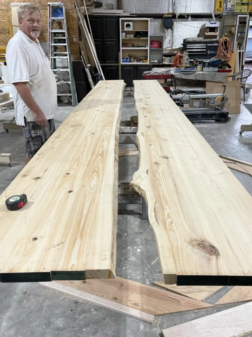 A man standing in a woodworking shop with two large, unfinished wooden slabs on a workbench.