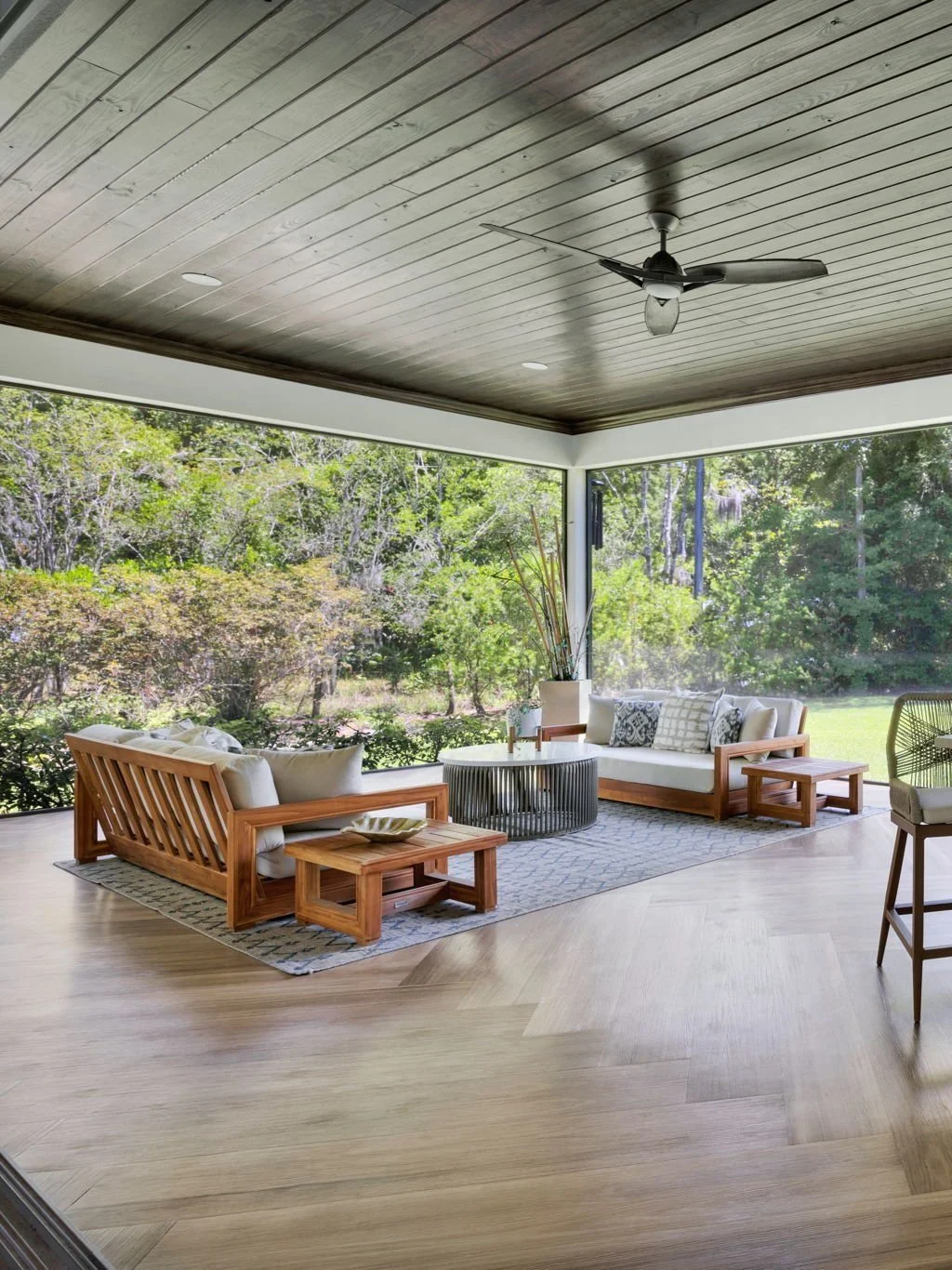 Living room with wooden furniture and large window overlooking trees and greenery, ceiling fan, light wood floor, and a decorative rug.